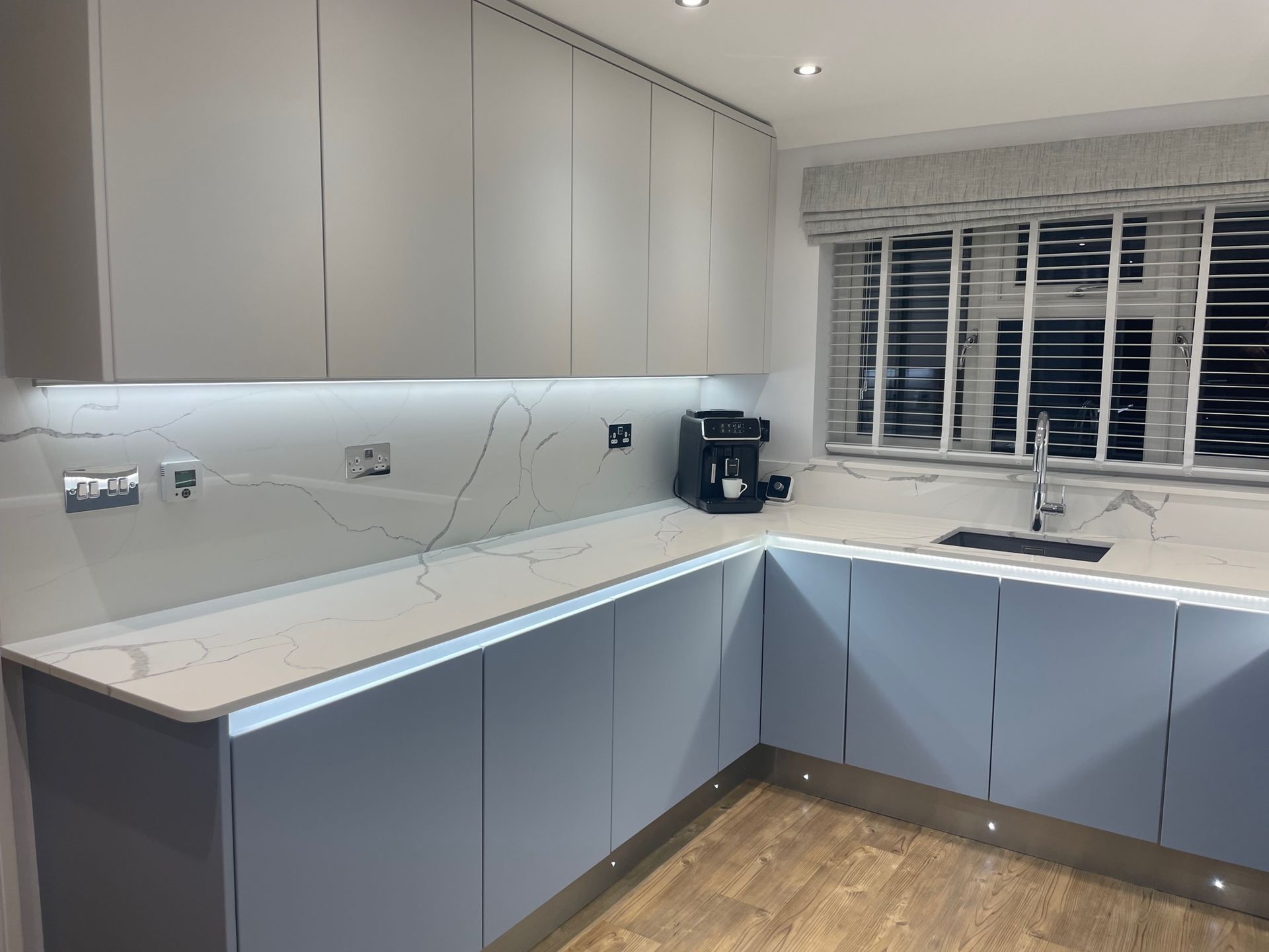 Modern kitchen with light blue base cabinets, cream wall cabinets, marble-effect counters, and under-cabinet lighting.