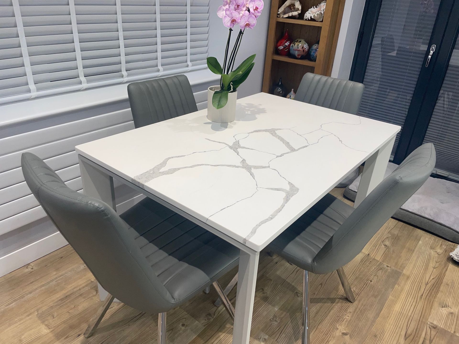 A white marble-effect dining table with four grey upholstered chairs in a room with wood flooring and blinds.