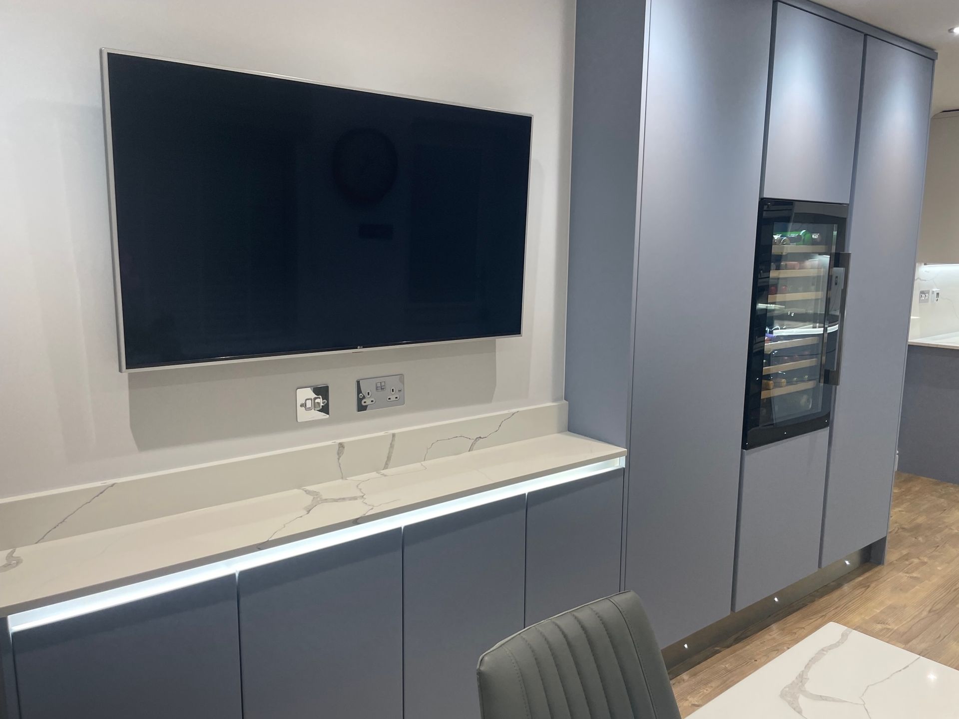 A flat-screen TV mounted above grey kitchen cabinets with a marble countertop, LED lighting, and an integrated wine cooler.
