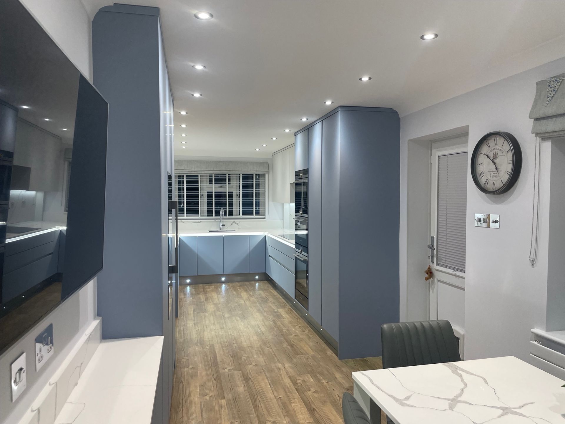 A modern kitchen with blue cabinetry, light wood floors, recessed lighting, and a dining area with a marble-topped table.