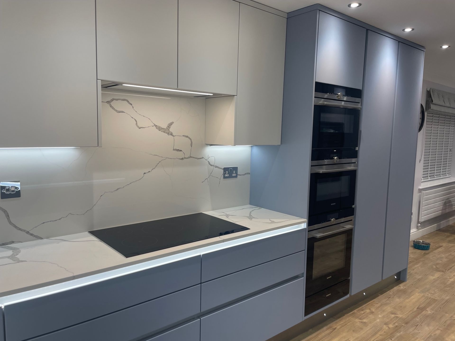 A modern kitchen featuring matte blue lower cabinets, off-white upper cabinets, a marble backsplash, and built-in appliances.