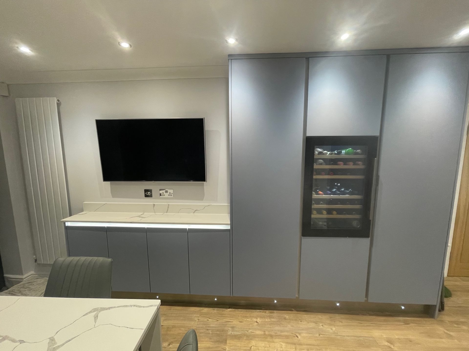Modern kitchen wall with gray cabinets, built-in wine cooler, wall-mounted television, and stone countertop with lighting.
