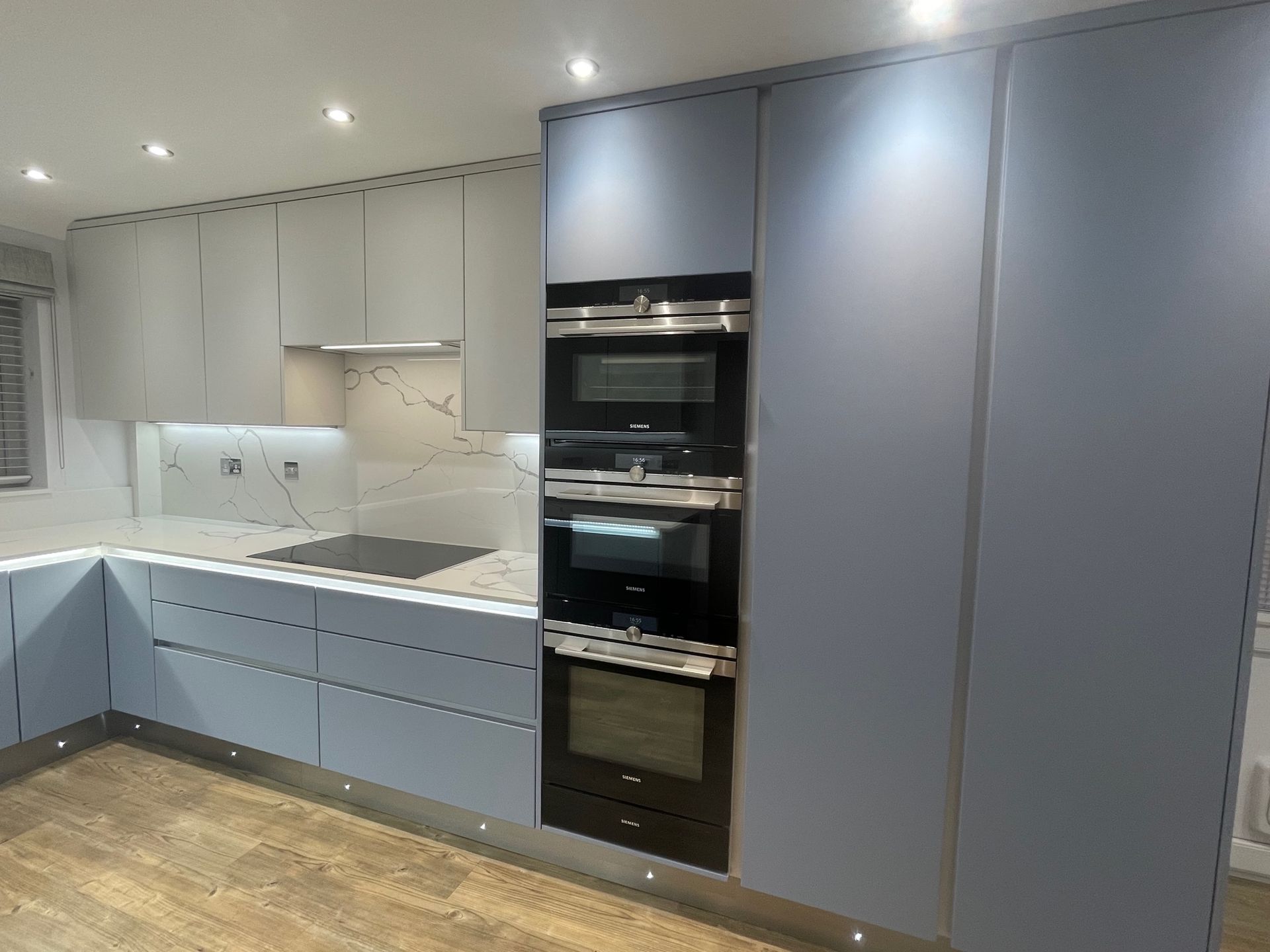 Modern kitchen featuring light grey cabinets, marble-patterned backsplash, wood-look flooring, and a built-in double oven.