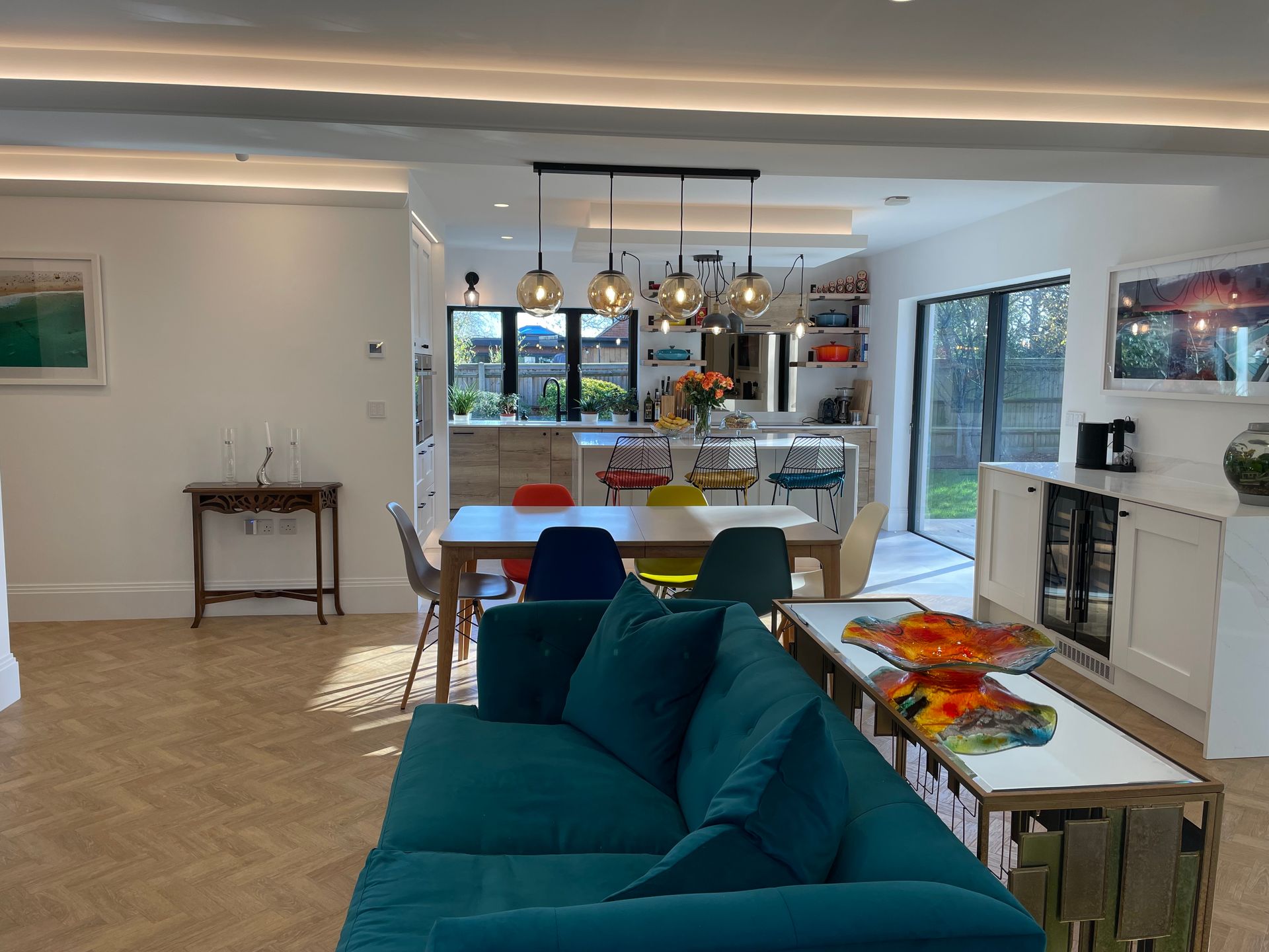 Modern, open-plan living room with a teal sofa, wooden dining table, and kitchen area with pendant lights.