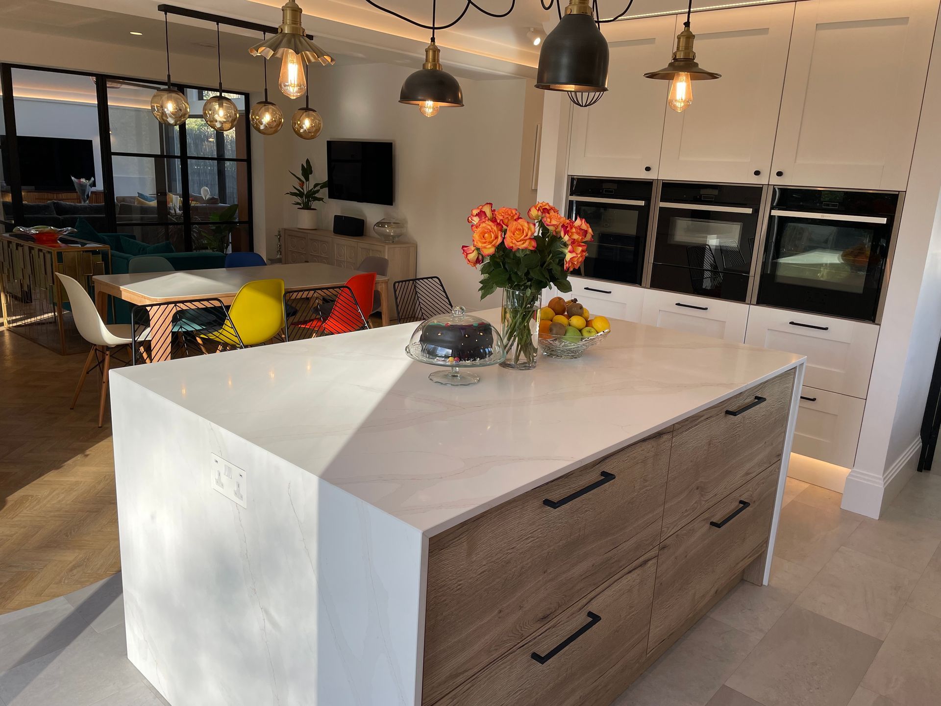 A modern kitchen with a large white quartz island, wood-grain cabinets, three built-in ovens, and hanging pendant lights.