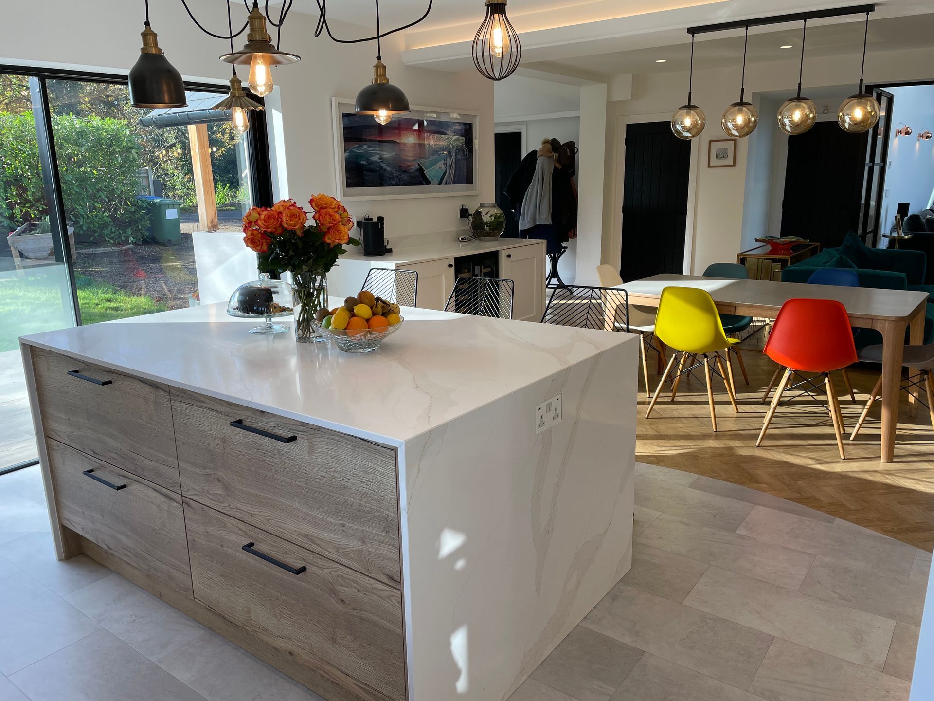 Modern kitchen with a large marble-topped island, wooden cabinetry, and bright red and yellow chairs at a dining table.