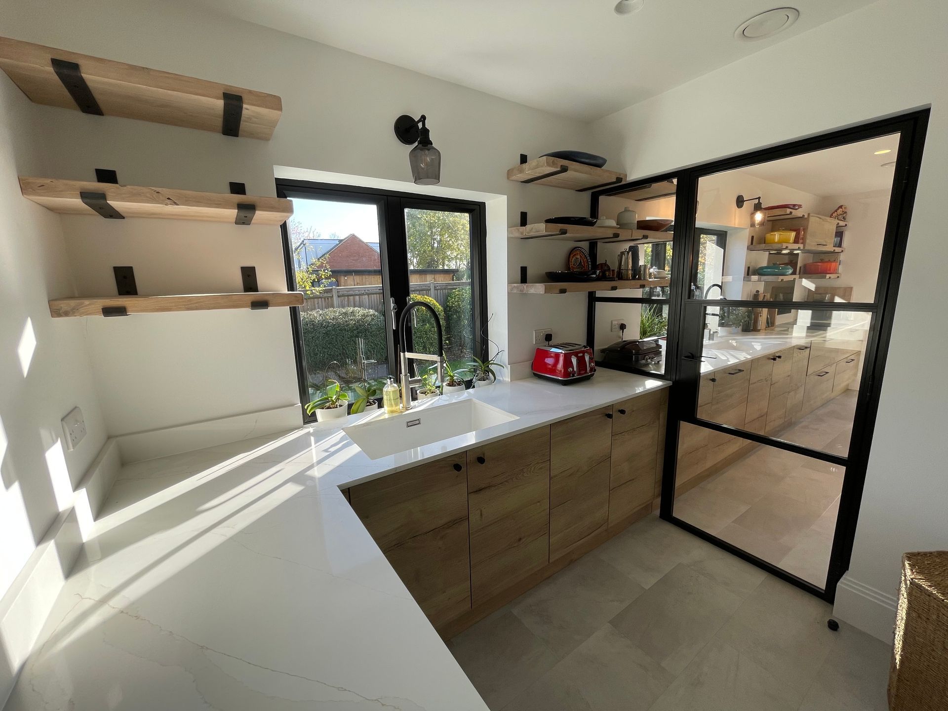 Modern kitchen with white countertops, wooden cabinets, floating shelves, and a large black-framed glass wall partition.