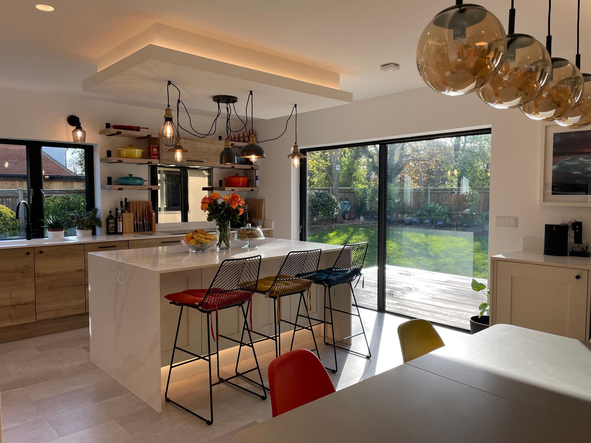 A bright, modern kitchen featuring a white island with colorful bar stools, open shelving, and a glass-walled garden view.
