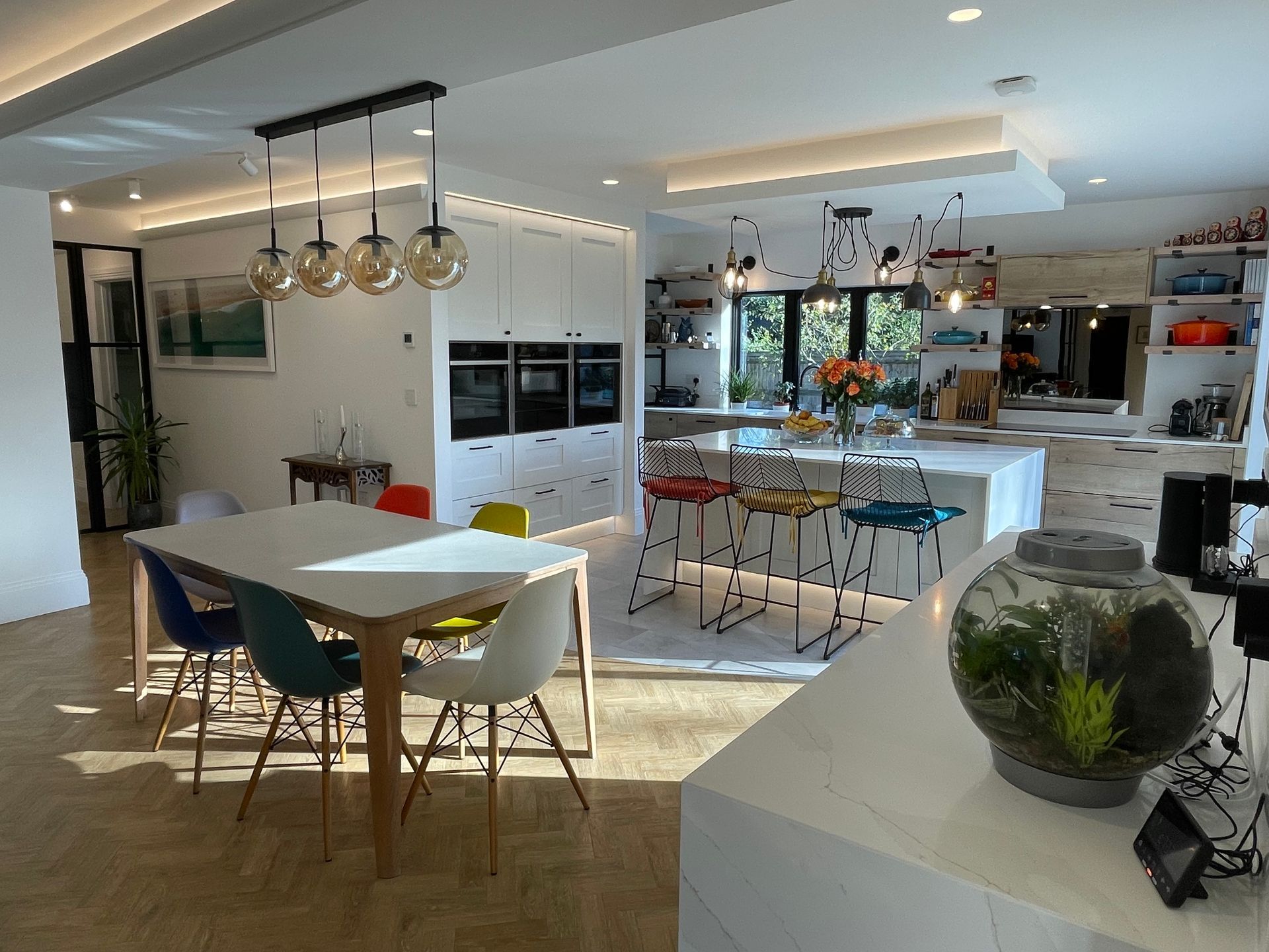 A modern open-plan kitchen with white cabinets, a dining table with colorful chairs, and a kitchen island with stools.