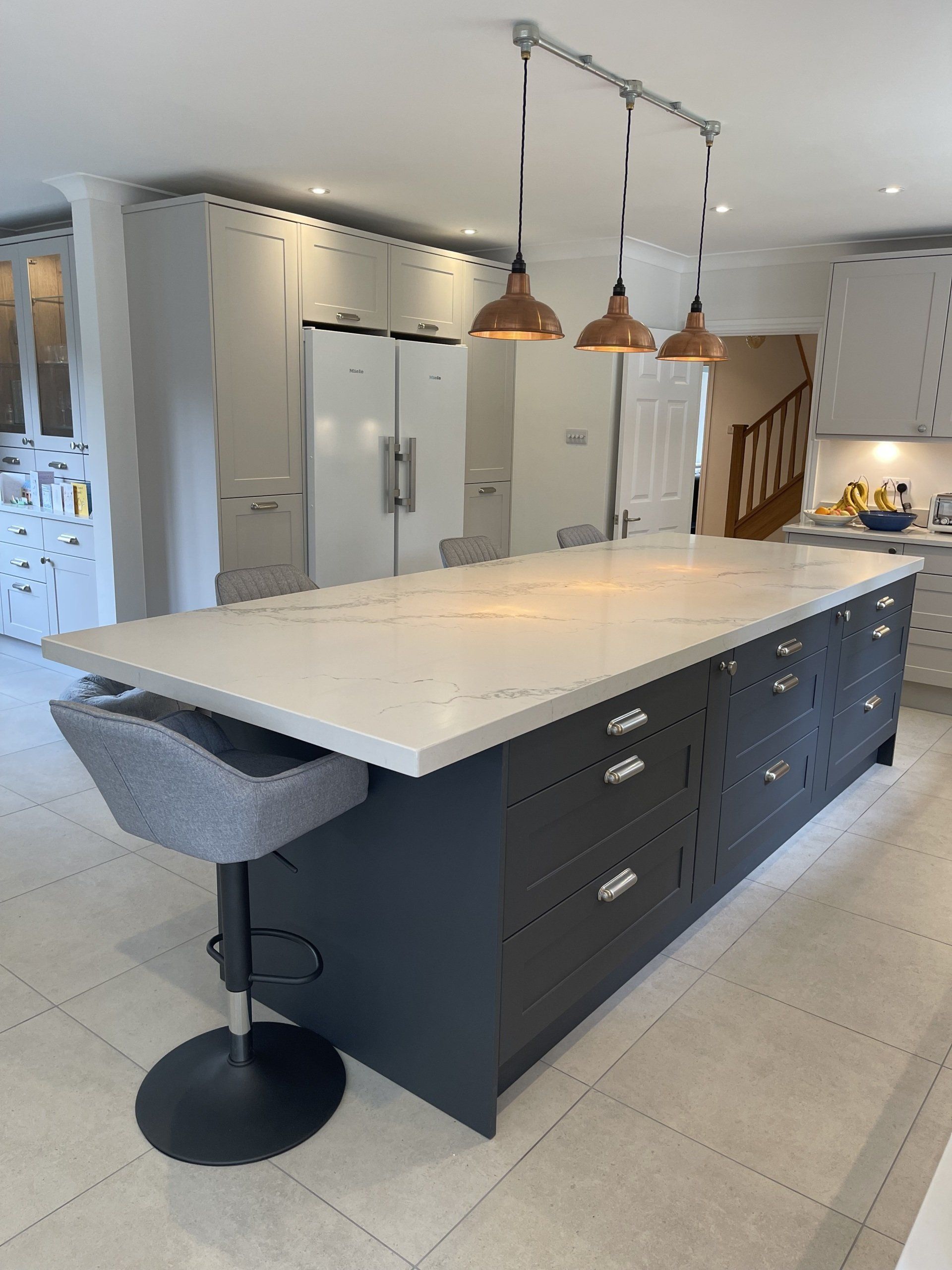 A kitchen island with a white countertop and charcoal-colored cabinets, featuring a single gray stool and overhead lights.