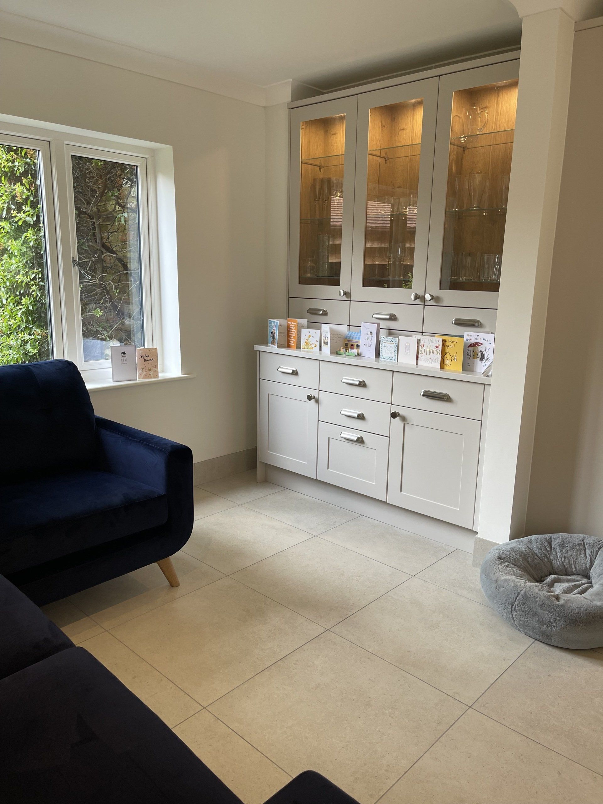 A bright room with a navy armchair, tiled floor, a large grey hutch filled with cards, and a pet bed in the corner.