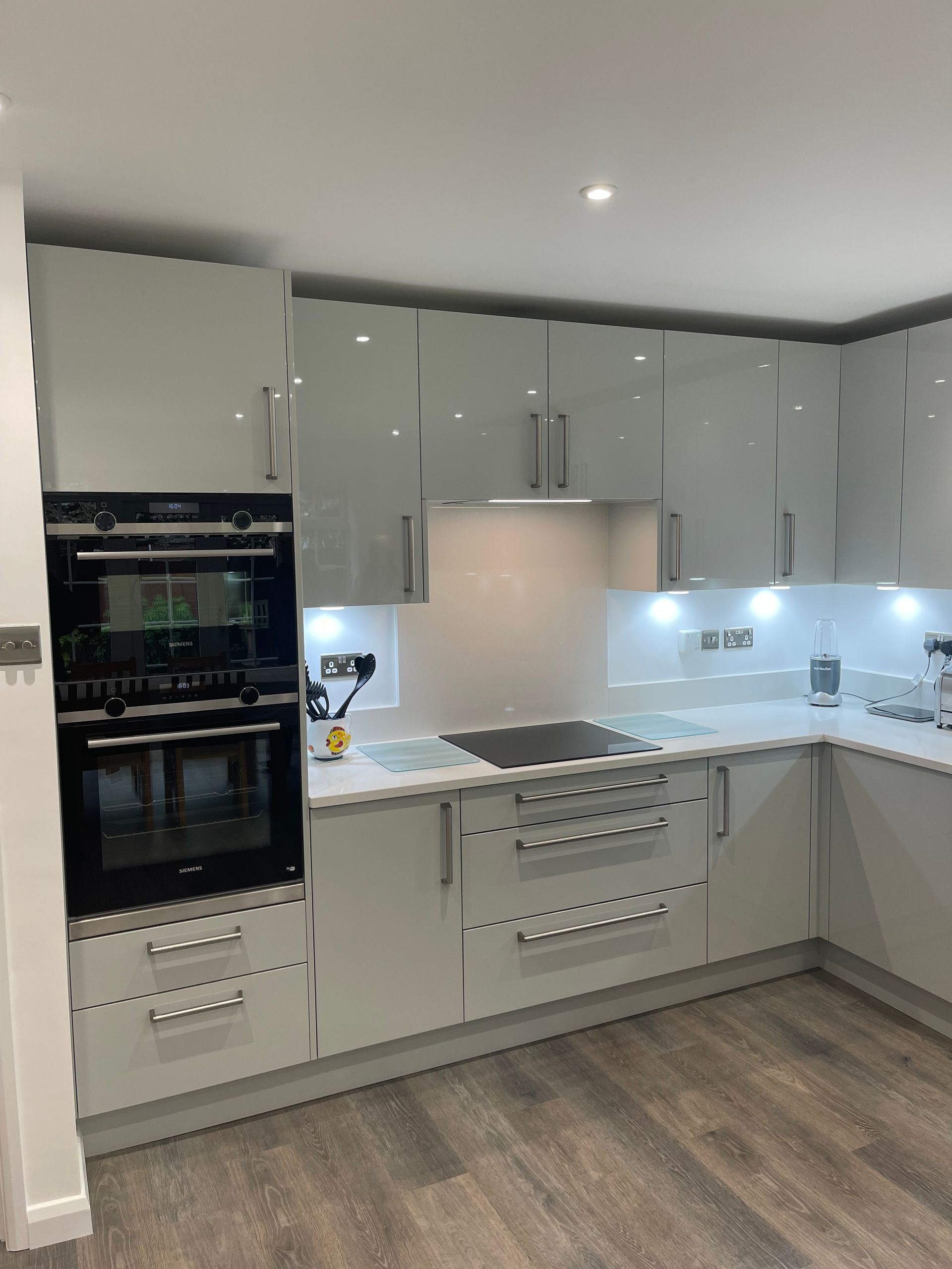 A modern kitchen with light grey cabinets, dark countertops, built-in double ovens, and under-cabinet LED lighting.