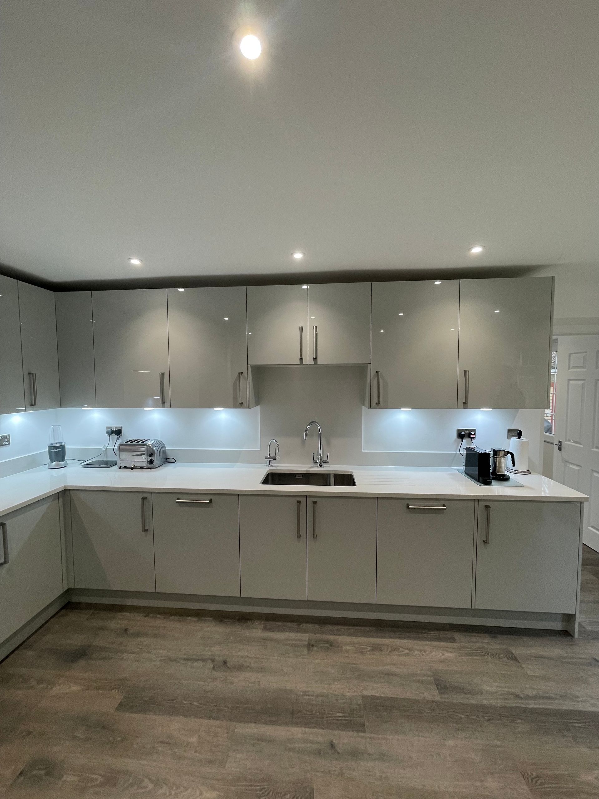 A modern kitchen featuring glossy light grey cabinets, white countertops, and under-cabinet lighting above a wood floor.