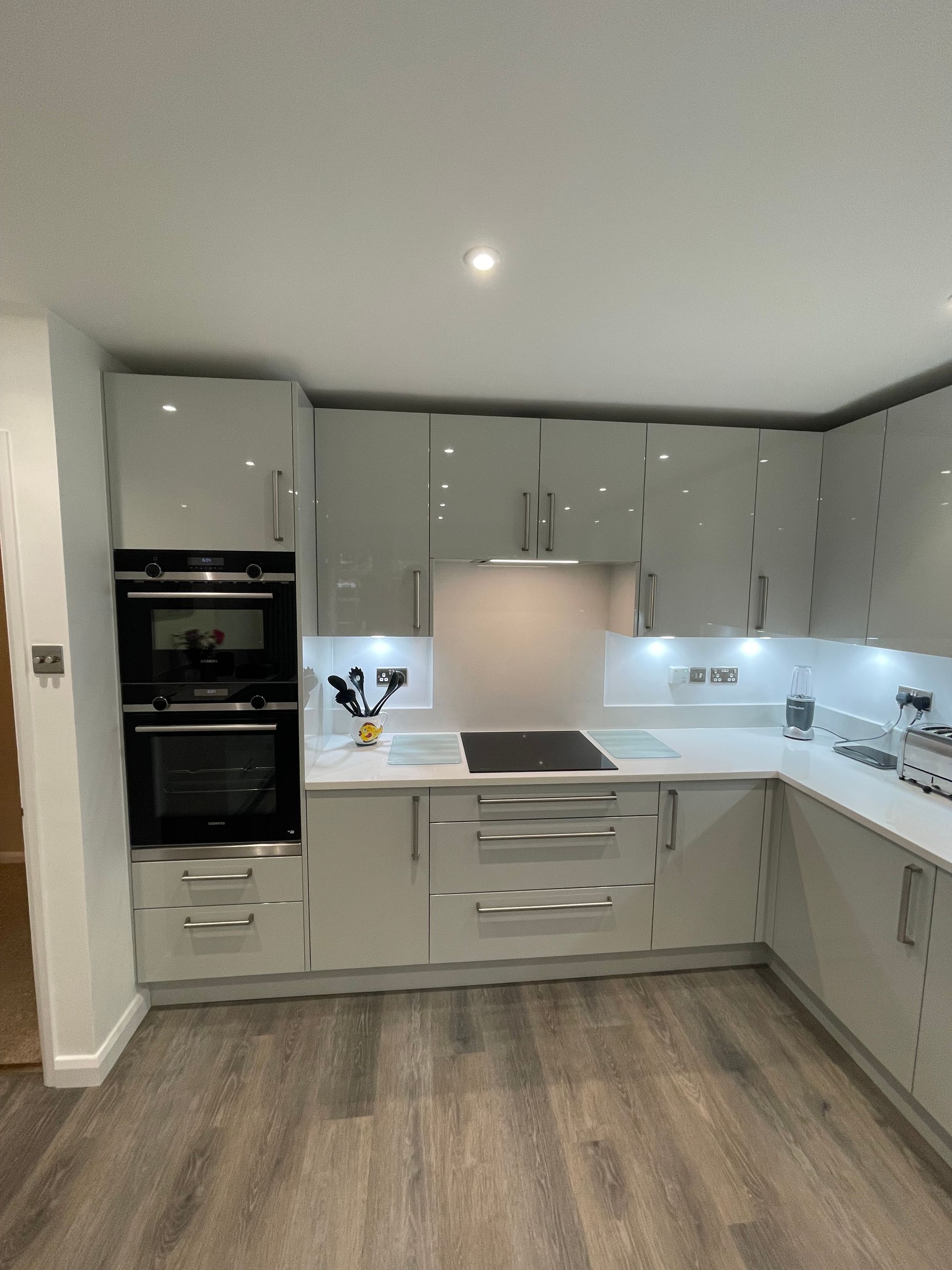 Modern kitchen with glossy grey cabinets, white countertops, built-in double oven, and wood-look flooring.