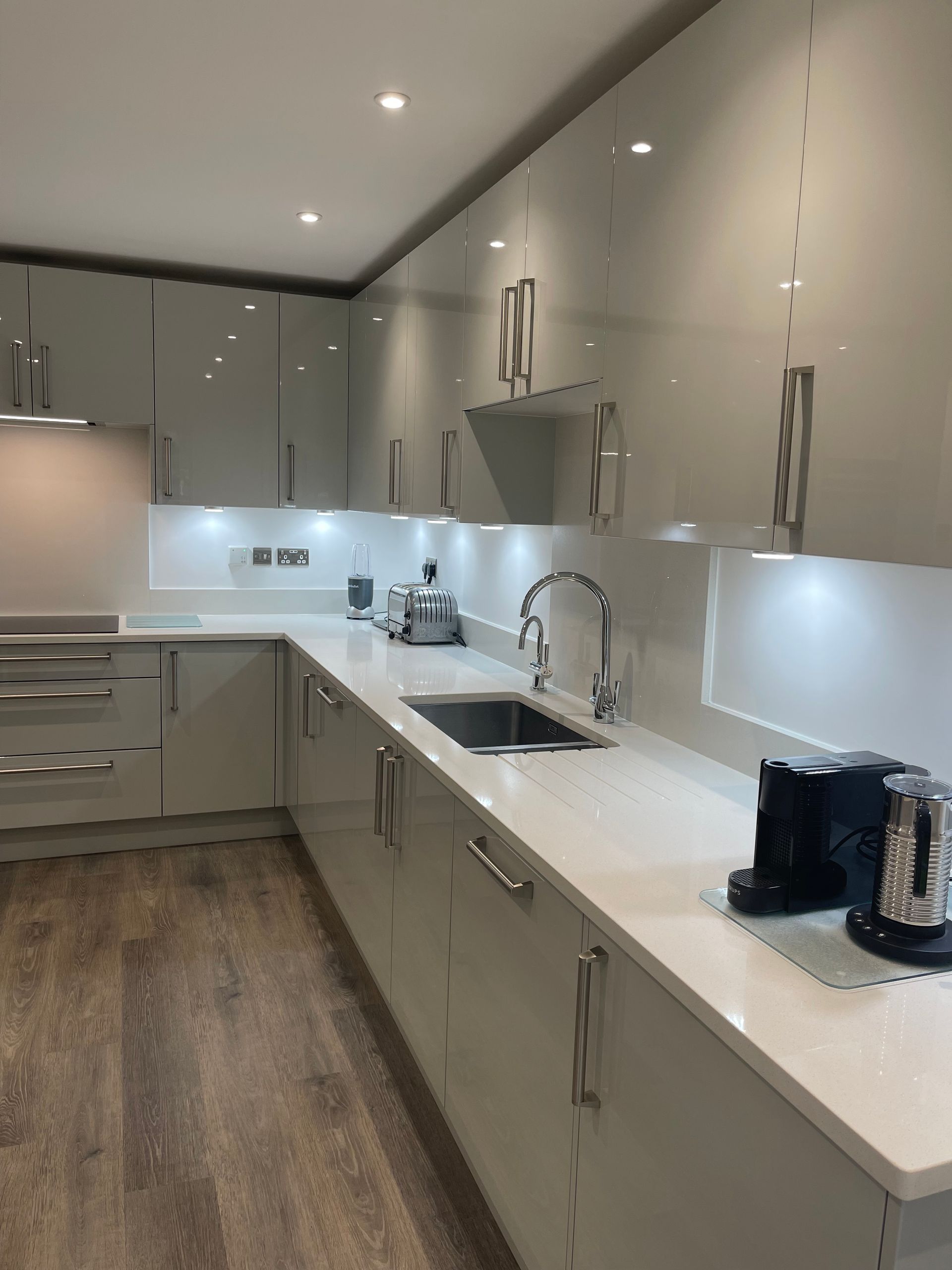 Modern kitchen with glossy light grey cabinets, white countertops, stainless steel sink, and wood-patterned flooring.