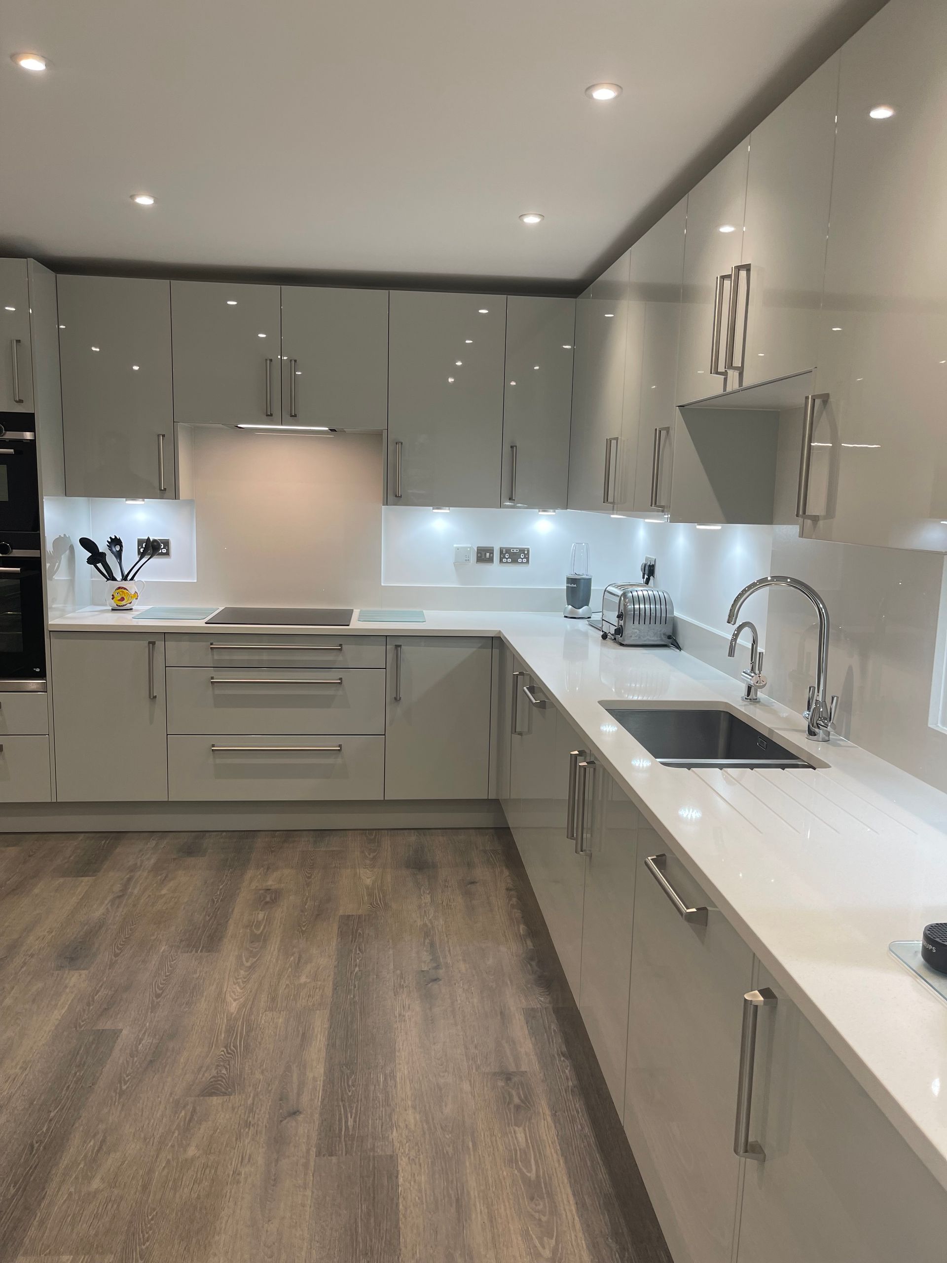 Modern kitchen with glossy light-grey cabinets, white countertops, stainless steel appliances, and wood-look flooring.