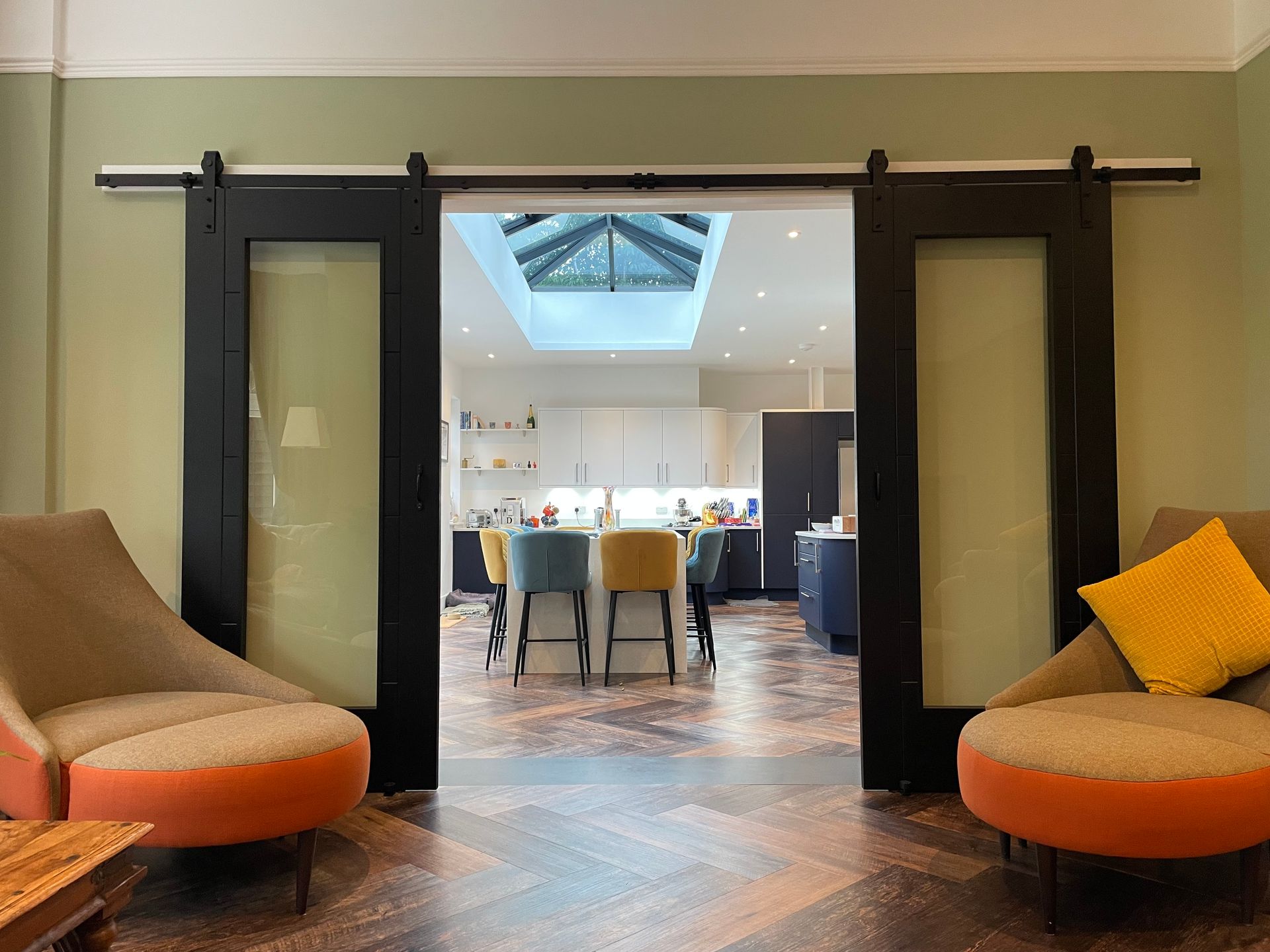 Black sliding barn doors open to a kitchen with a skylight and a central island, flanked by two orange and beige armchairs.