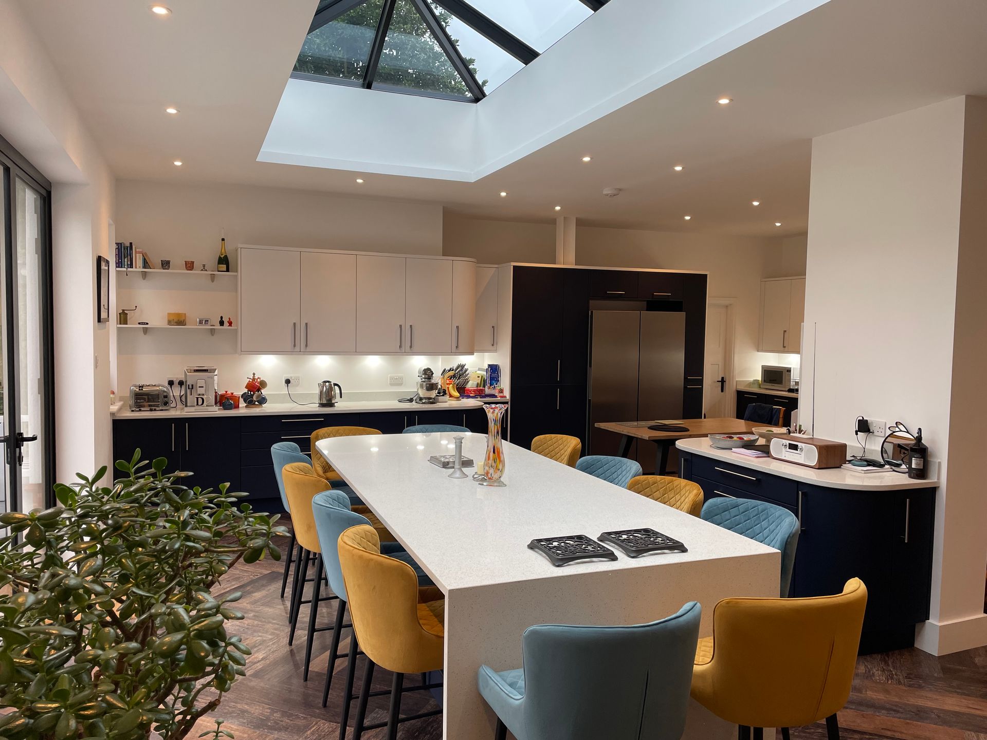 Modern kitchen with a long white island, yellow and blue bar stools, dark cabinets, and a large roof lantern skylight.