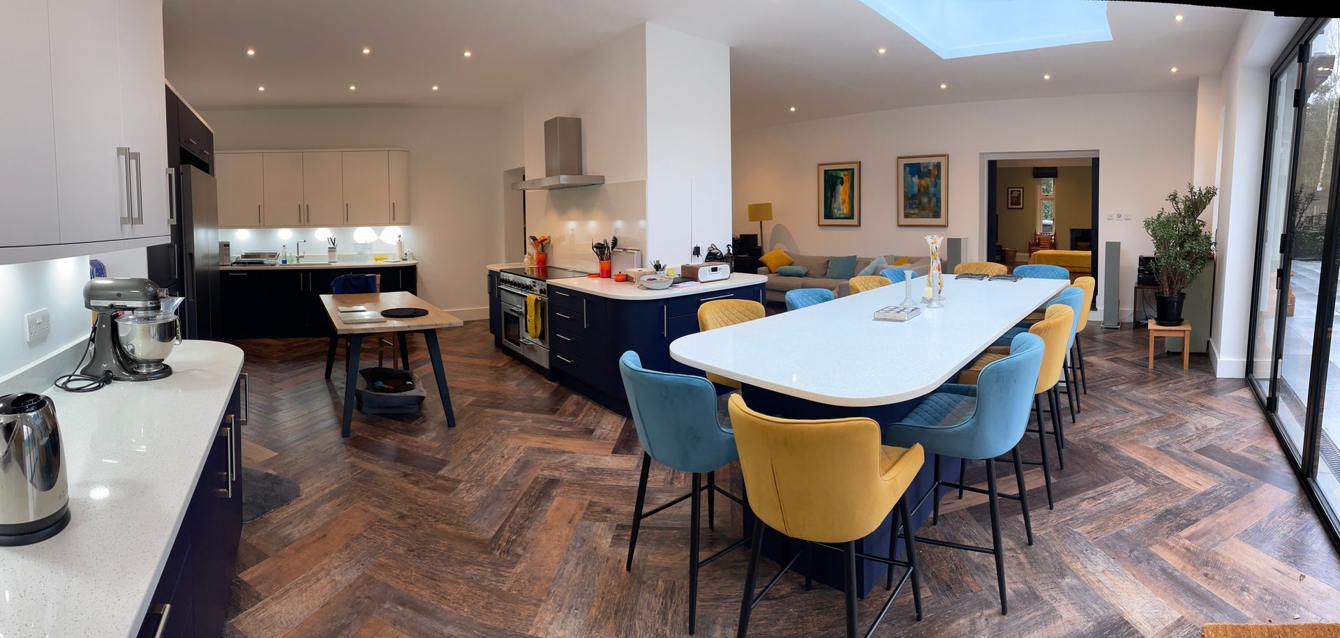 Modern, open-plan kitchen with a large central island, white counters, navy cabinets, and yellow and blue bar stools.