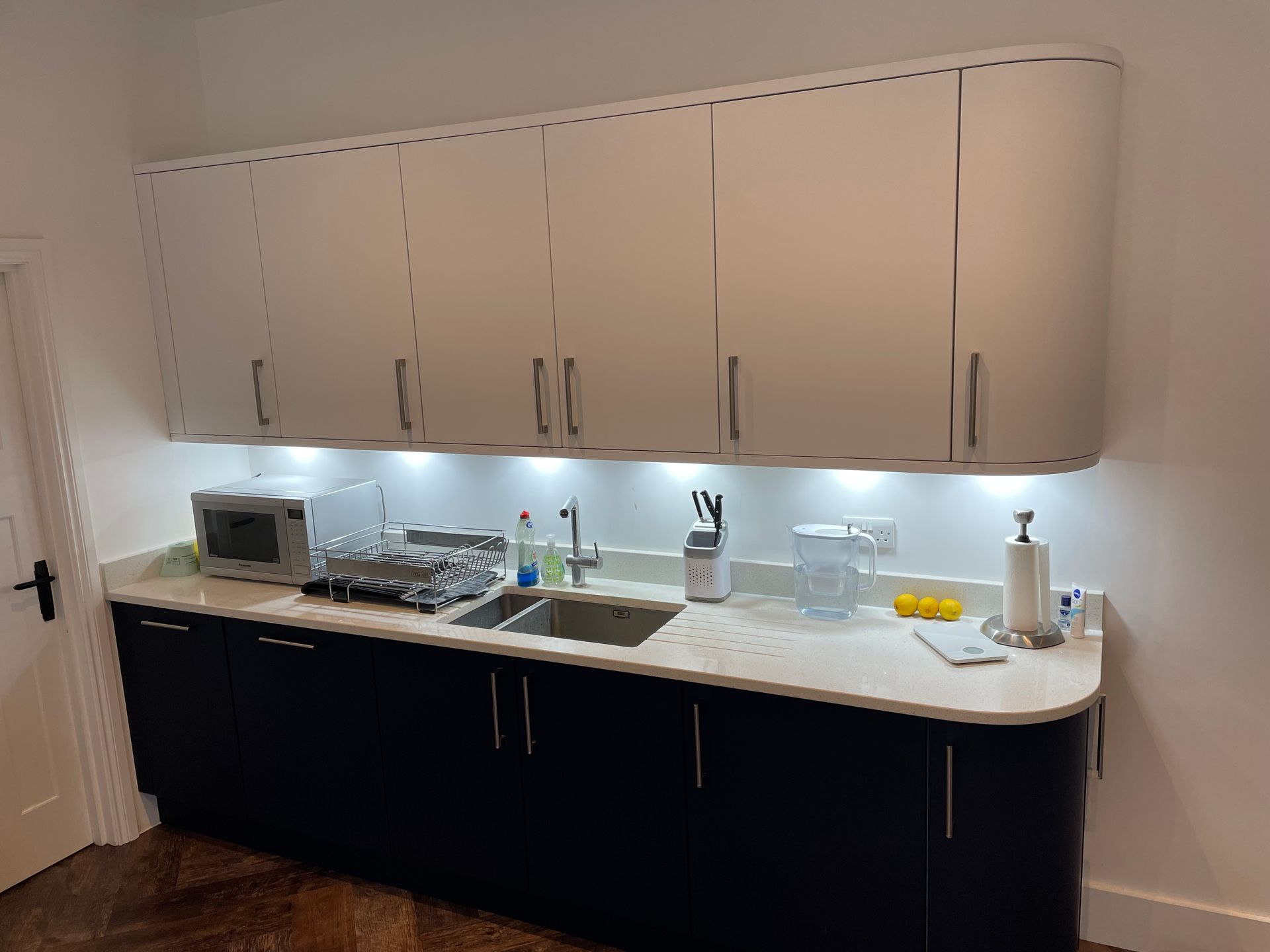 A modern kitchen kitchenette with white upper cabinets, dark lower cabinets, a light countertop, sink, and under-cabinet LED.