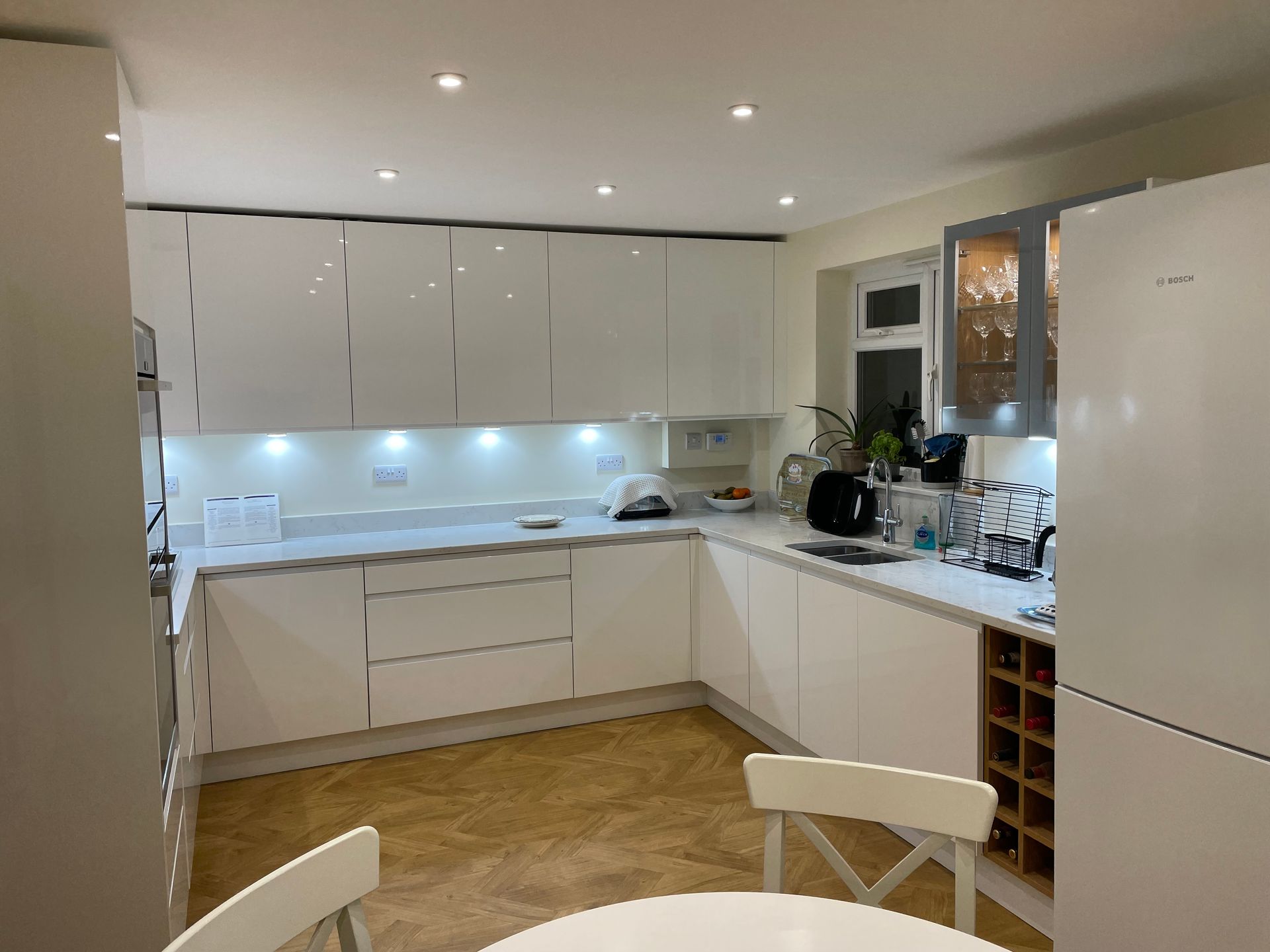 A modern U-shaped kitchen with white gloss cabinets, light-colored countertops, under-cabinet lighting, and tile floors.