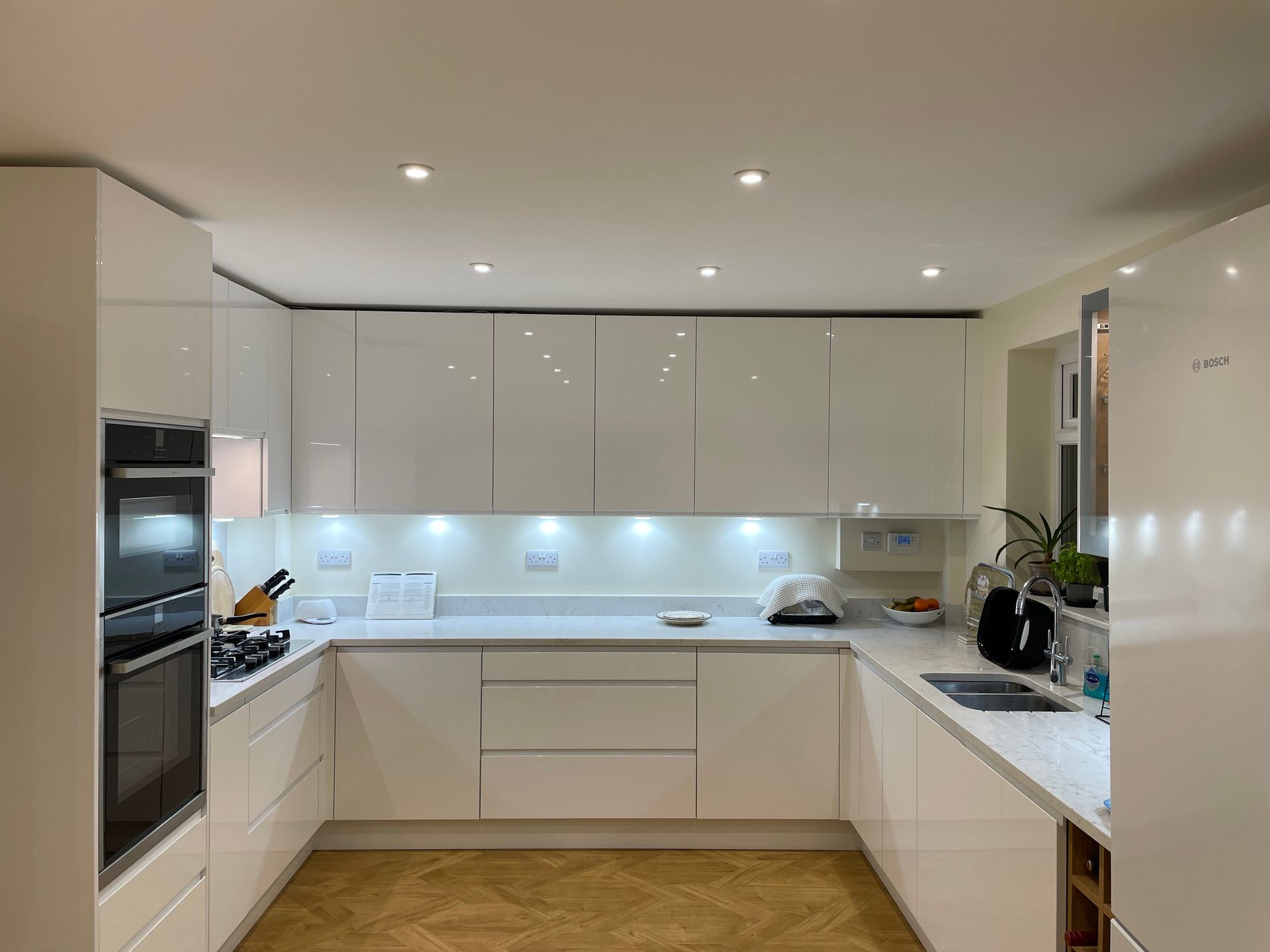 A modern U-shaped kitchen with white gloss cabinets, light countertops, integrated appliances, and warm floor lighting.
