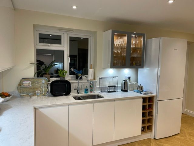 A bright kitchen with white cabinets, a stainless steel sink, a glass-front wall cabinet, and a white refrigerator.