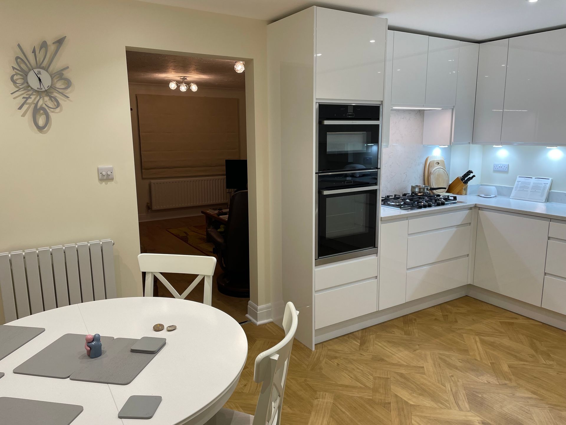 A modern kitchen with white cabinetry, a built-in double oven, a round dining table, and a view into a living room.