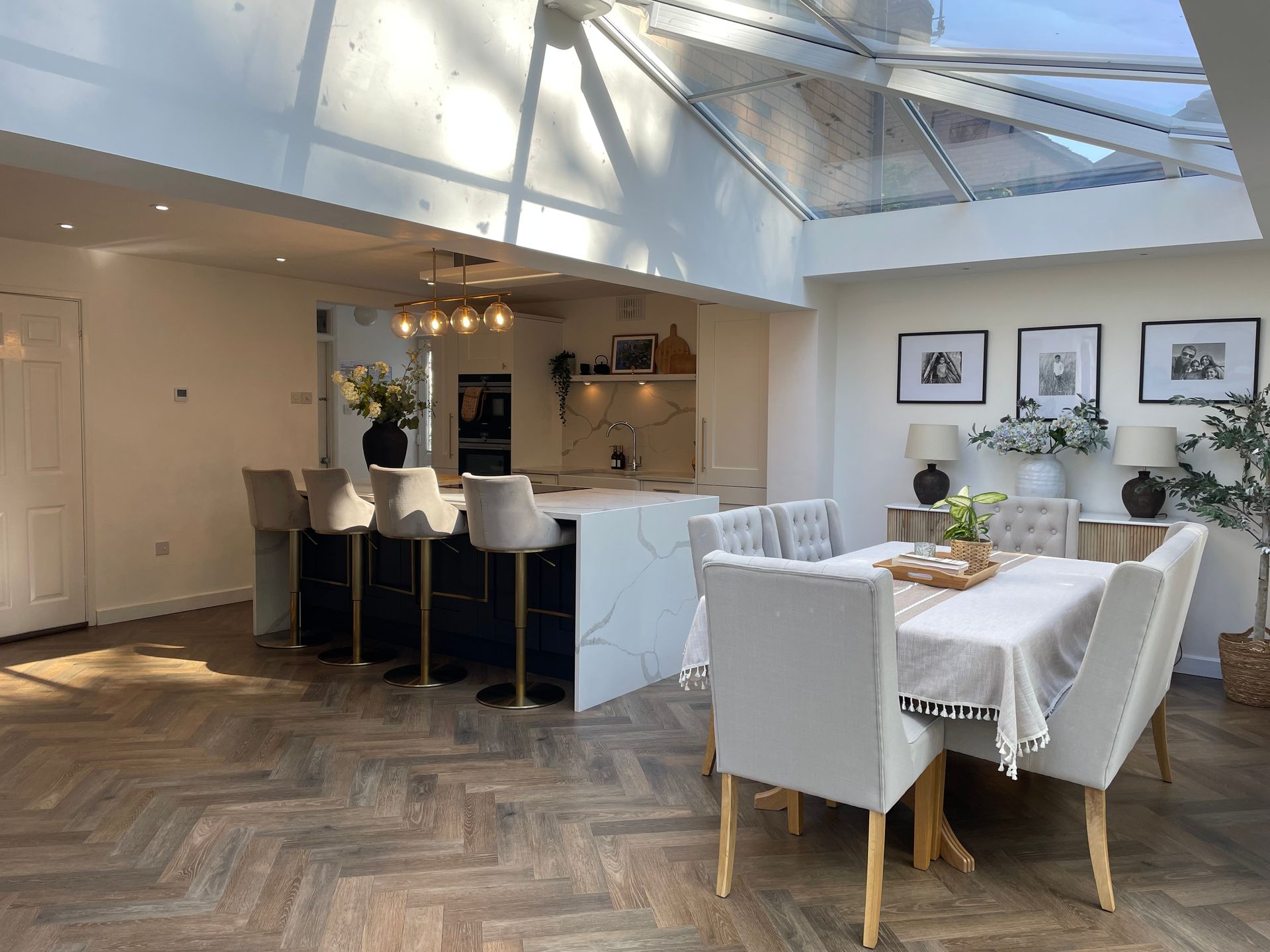 A modern open-plan kitchen and dining area with a glass ceiling, herringbone floors, a kitchen island, and light furniture.