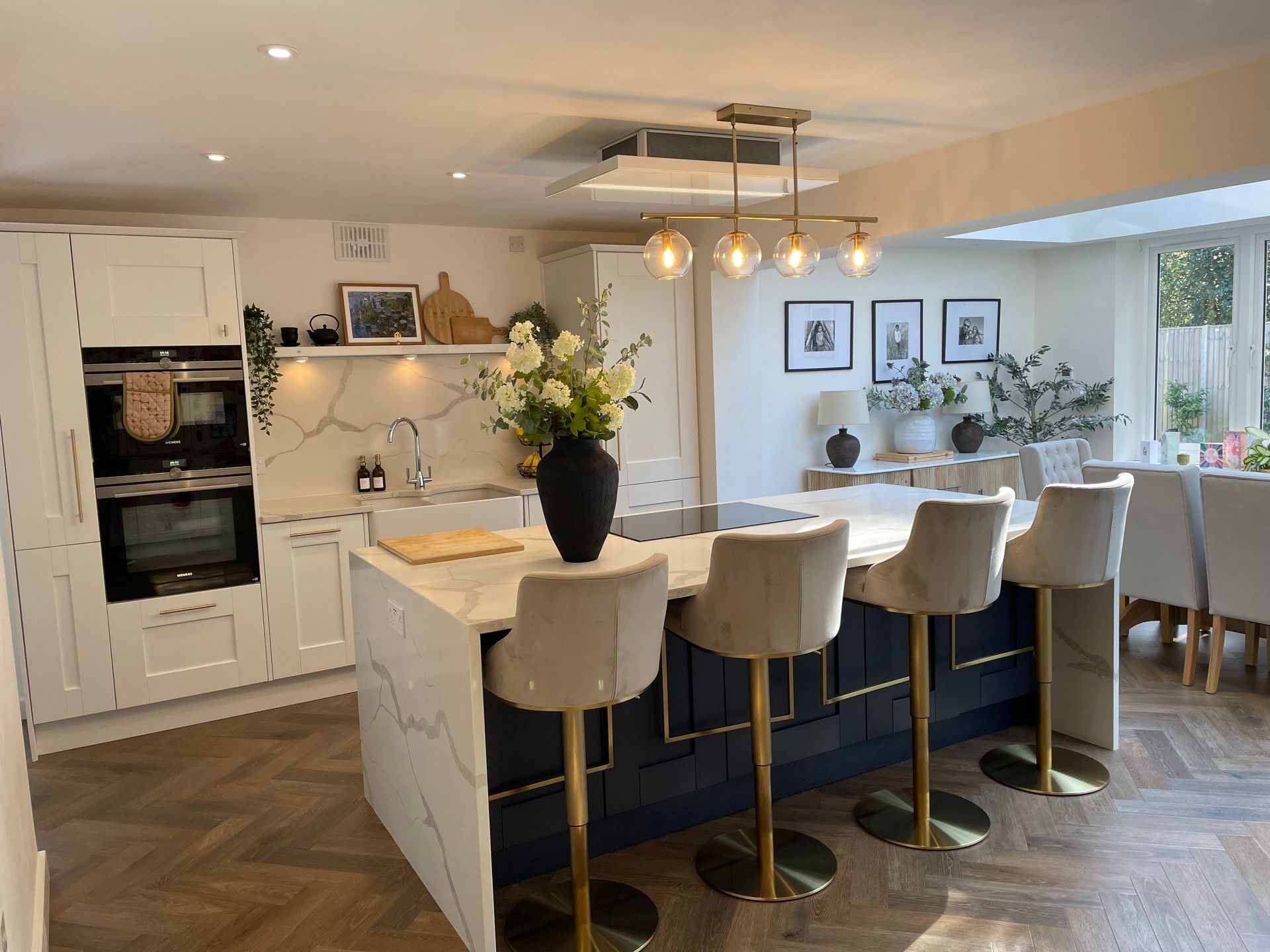 Modern kitchen with a dark blue island, white cabinets, marble countertops, four bar stools, and herringbone flooring.