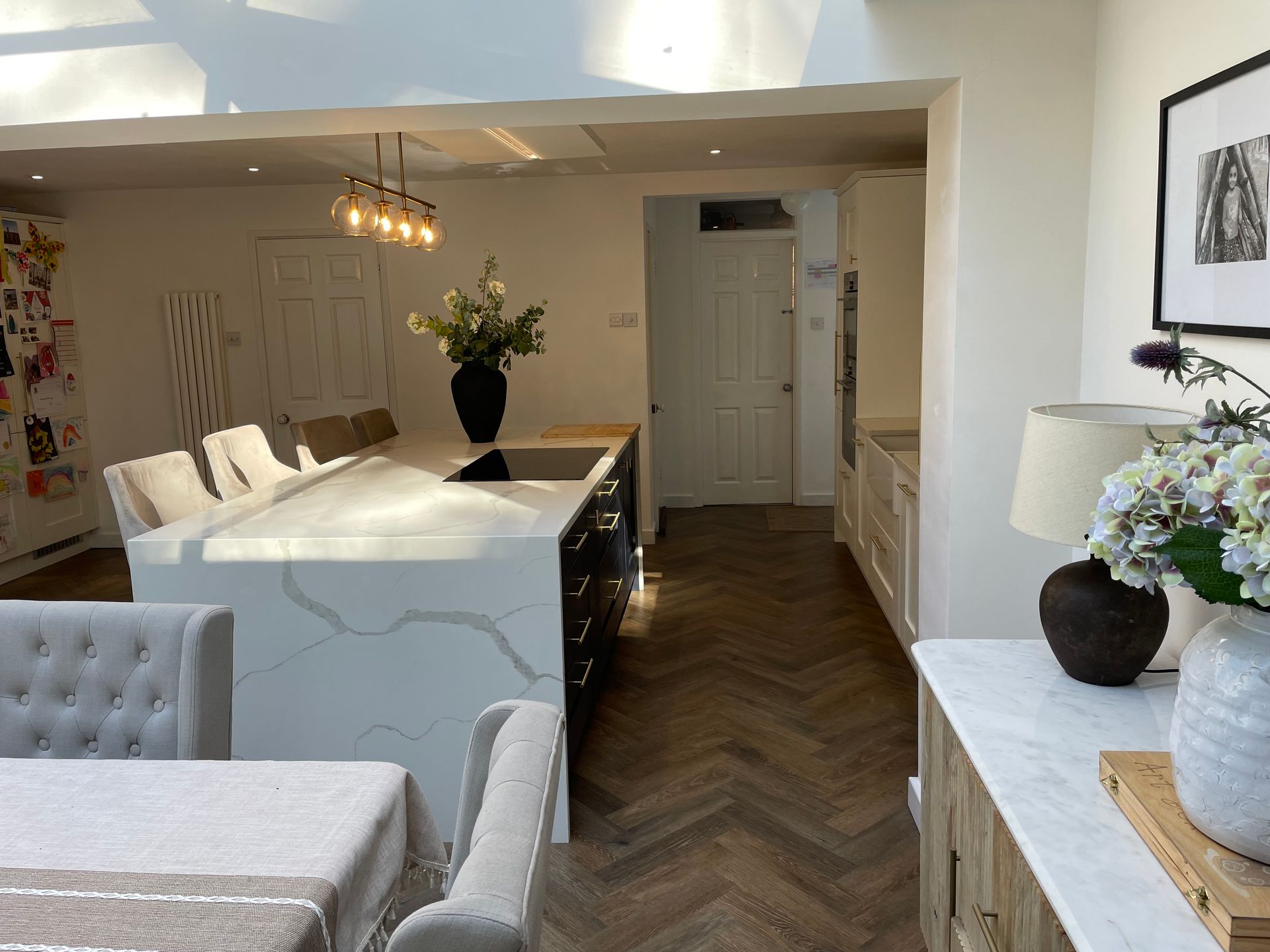 A modern kitchen with a large white marble-topped island, dark cabinetry, and herringbone wood flooring.