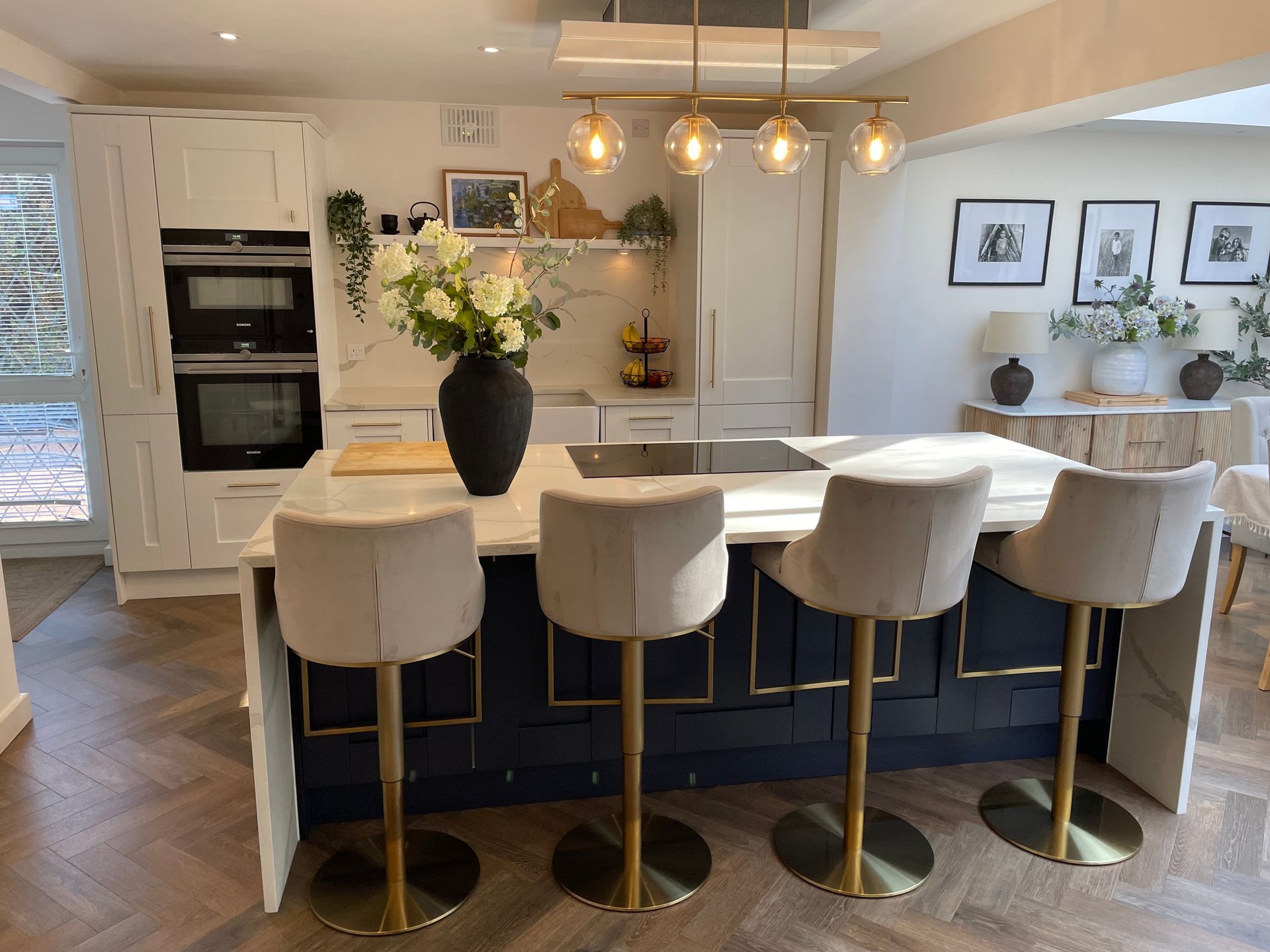 A modern kitchen featuring a dark blue island with four beige bar stools, a white marble countertop, and gold accents.