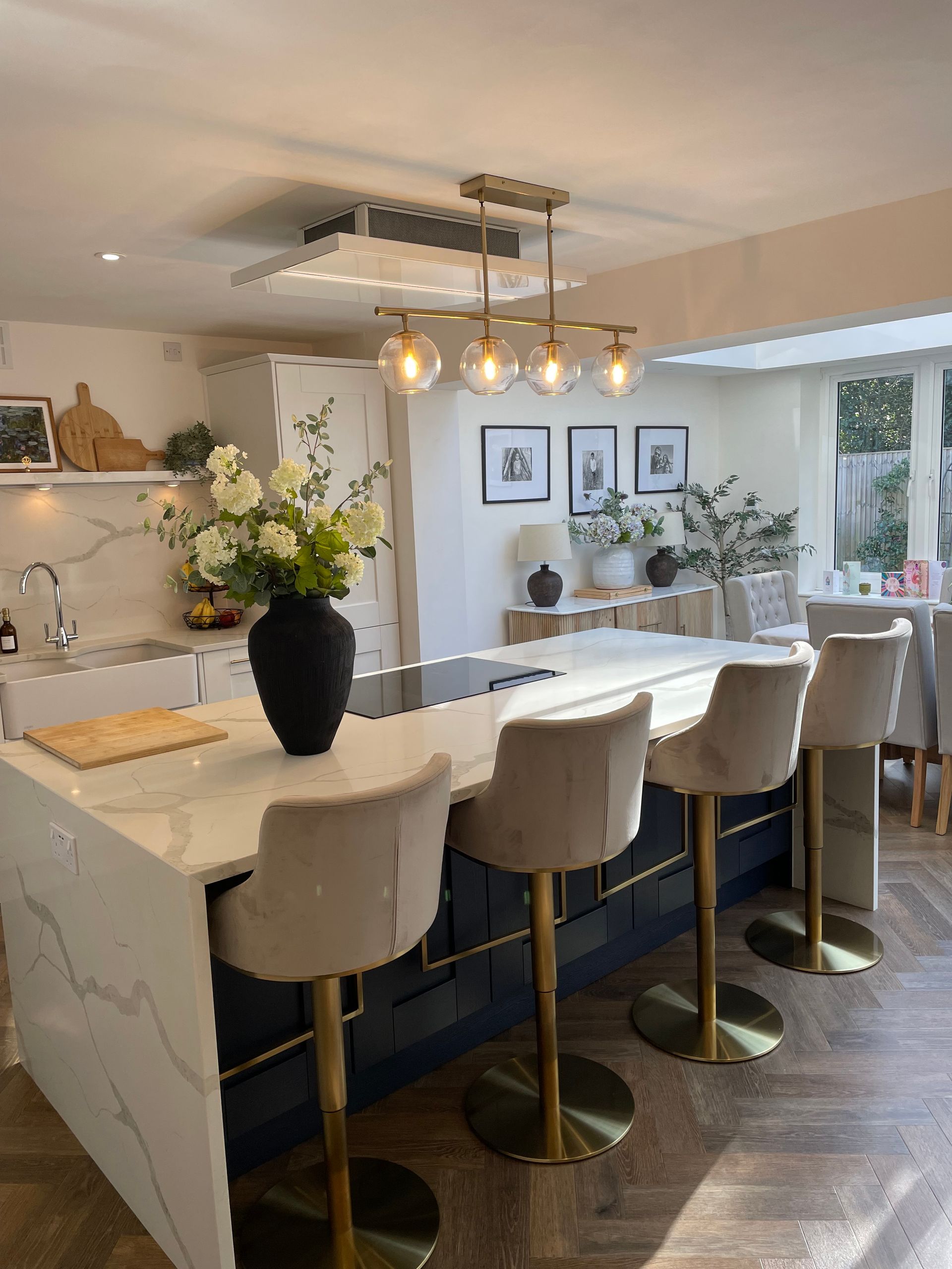 A modern kitchen island with four cream-colored velvet bar stools sits on a herringbone floor beneath a gold light fixture.