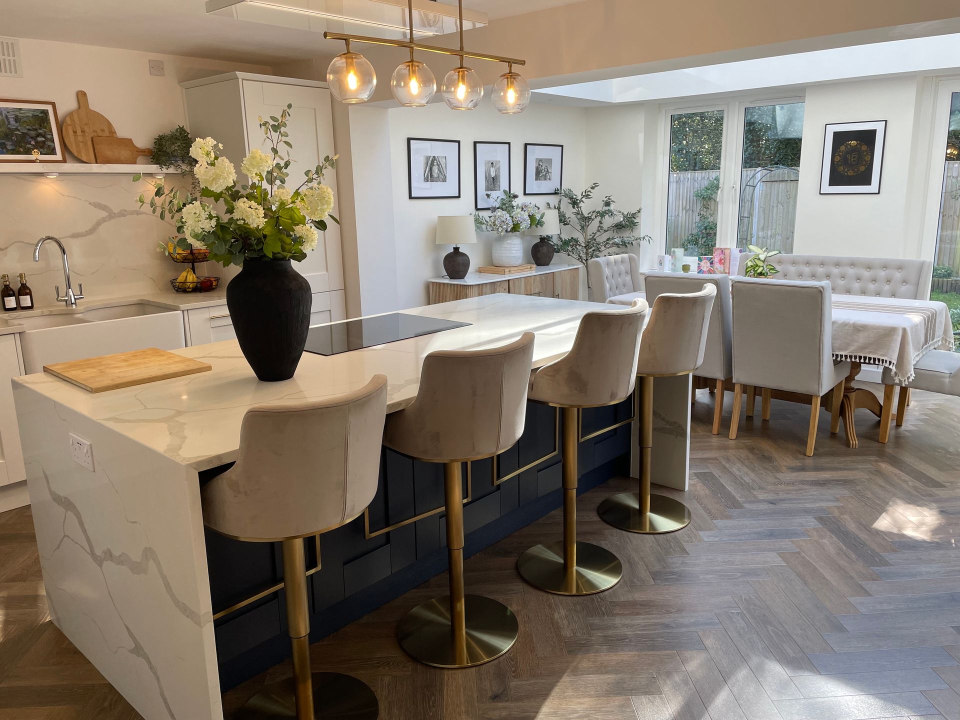 A bright, modern kitchen with a navy island, marble-style countertop, four velvet bar stools, and a dining area beyond.