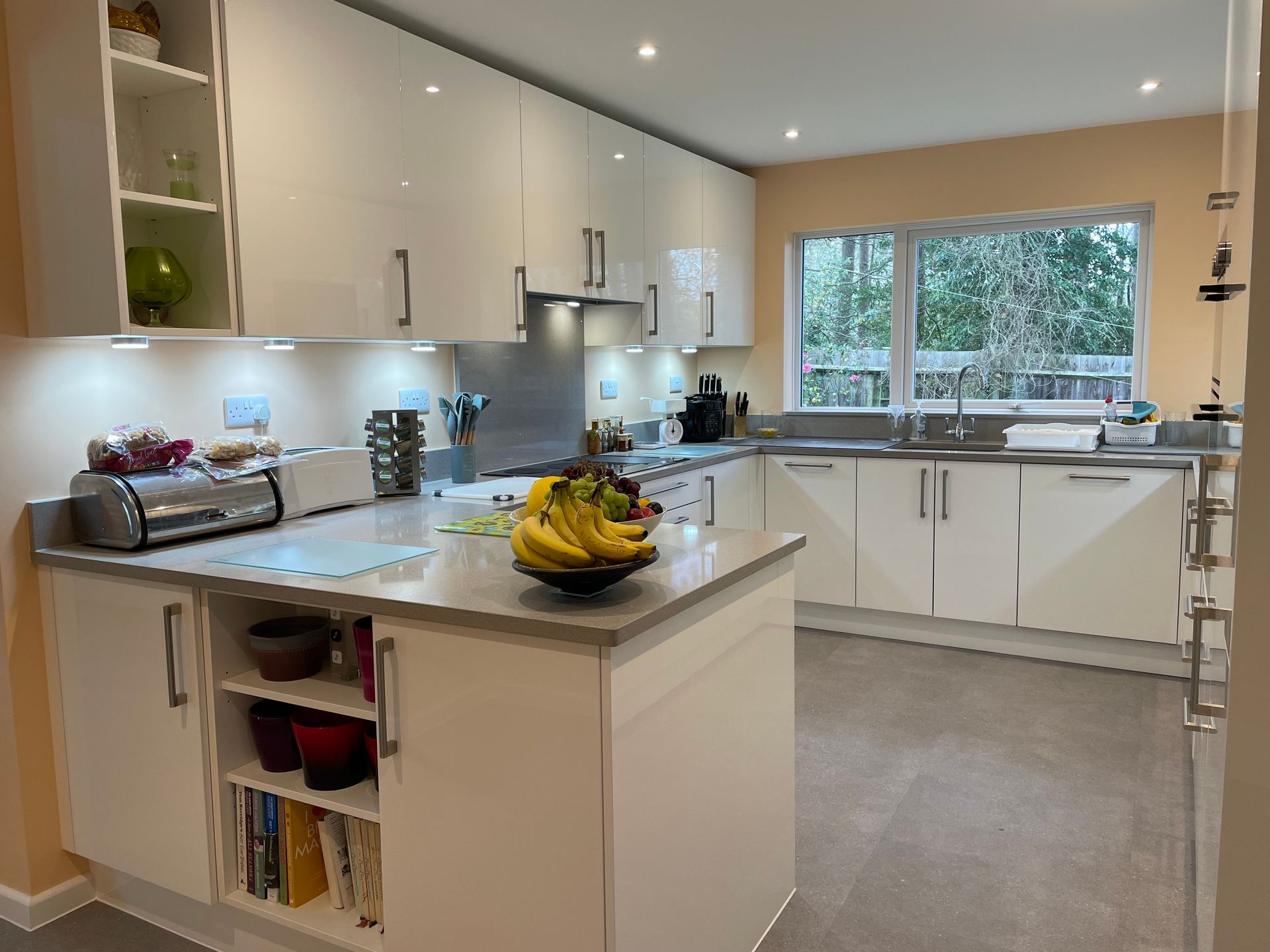 A modern kitchen with white cabinets, light countertops, a fruit bowl on the island, and a window overlooking greenery.