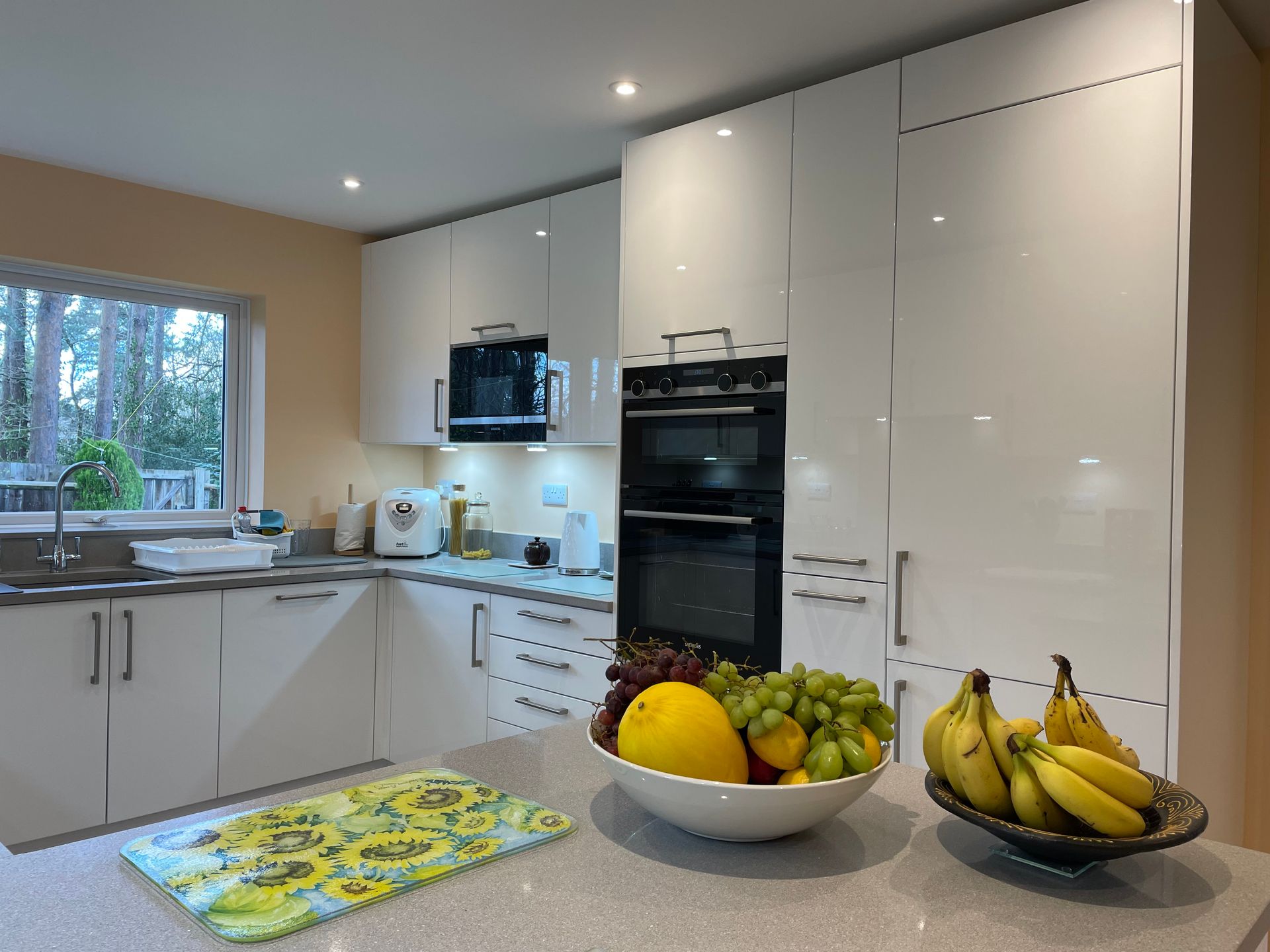 A modern kitchen with white glossy cabinets, a built-in oven, granite countertops, and two bowls of fresh fruit.