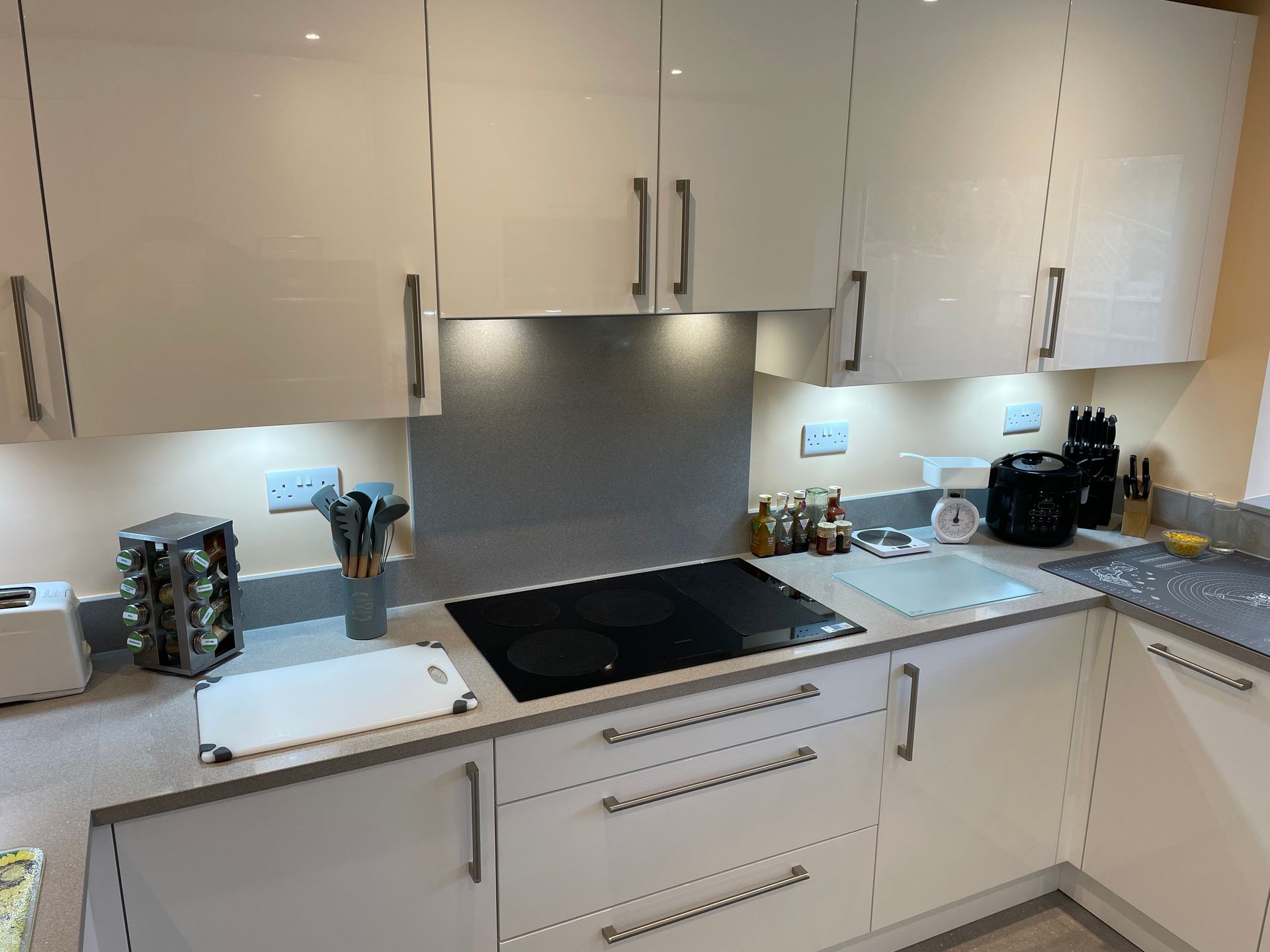 A modern kitchen with white glossy cabinets, a black electric stovetop, and grey countertops with under-cabinet lighting.