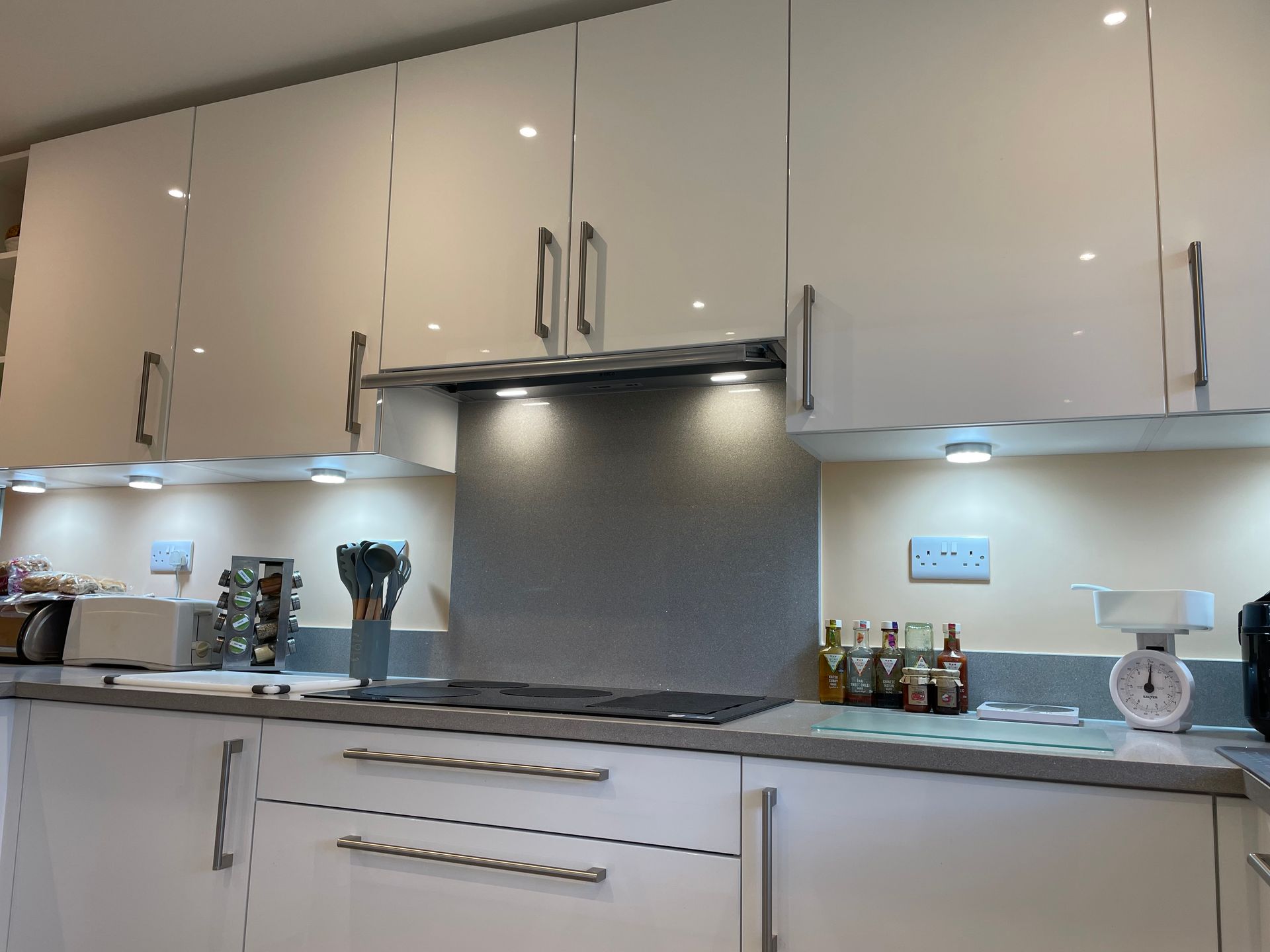 A bright modern kitchen with white gloss cabinets, a grey tiled backsplash, under-cabinet lighting, and kitchen appliances.