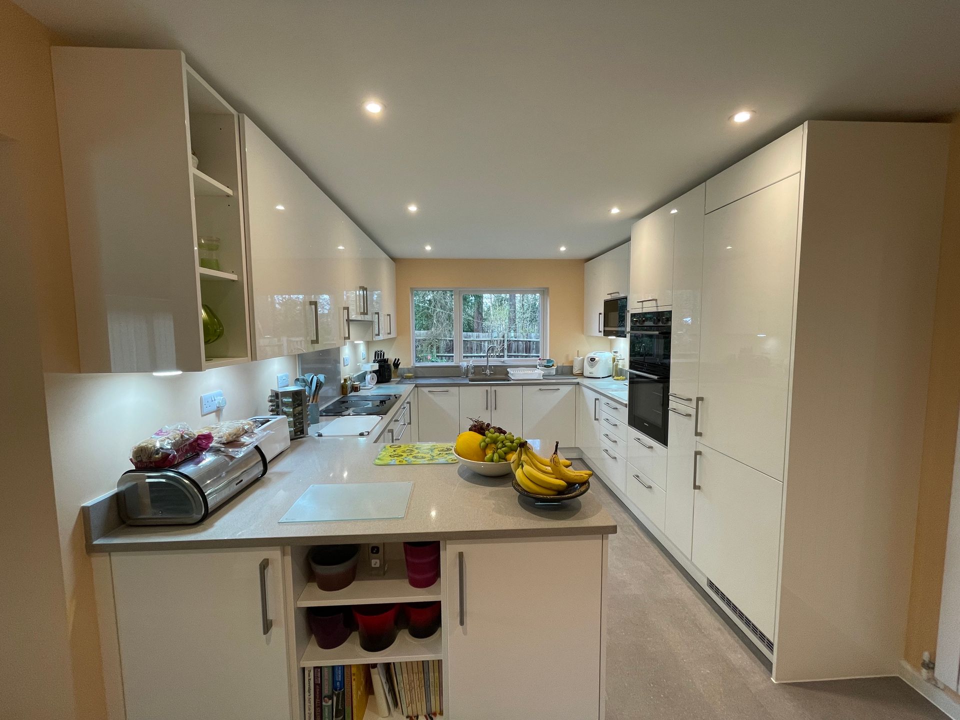 A modern white kitchen featuring an island, glossy cabinets, built-in appliances, and a window overlooking a garden.