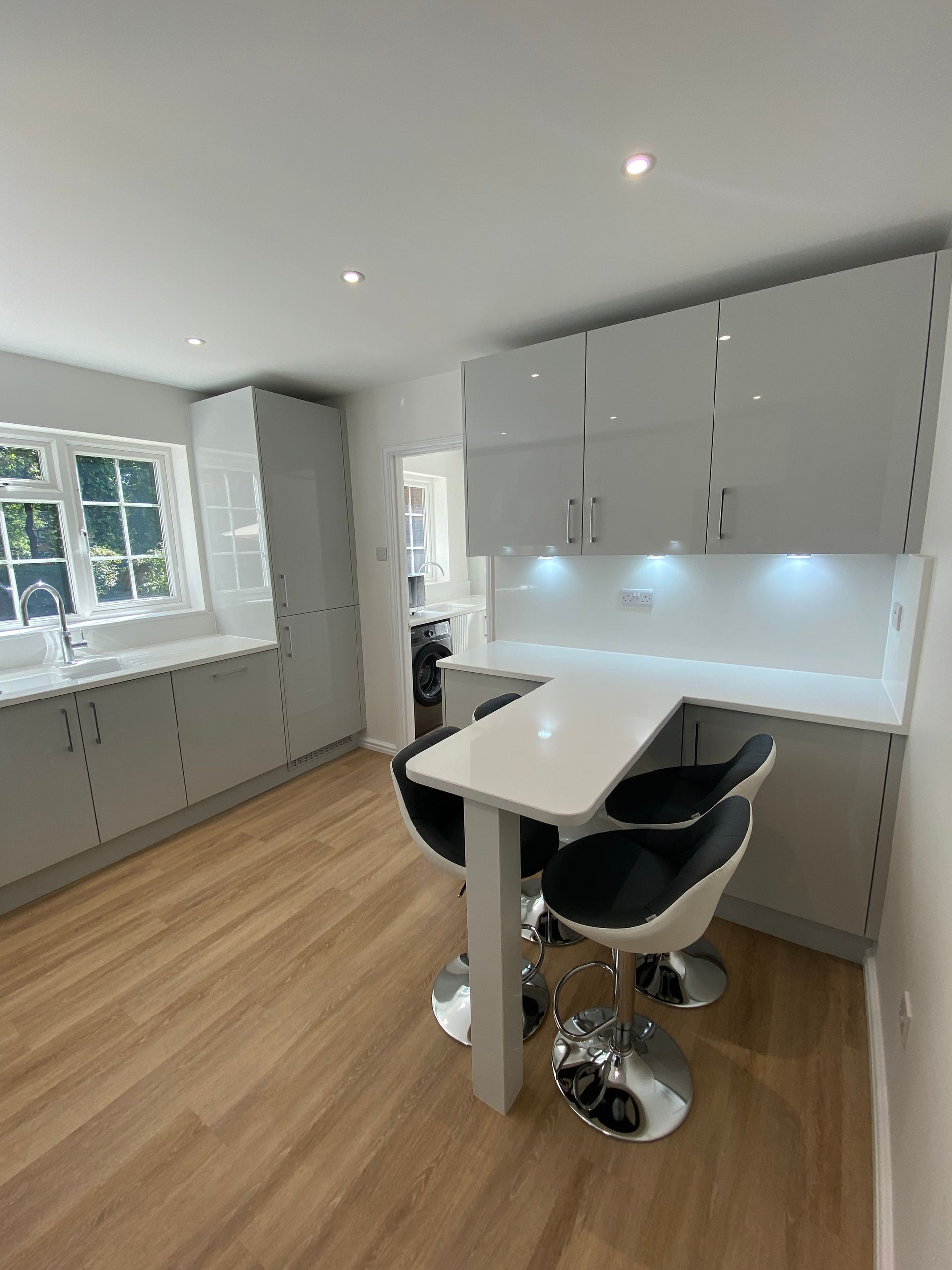 Modern kitchen with light grey glossy cabinets, white countertops, light wood flooring, and a breakfast bar with two stools.