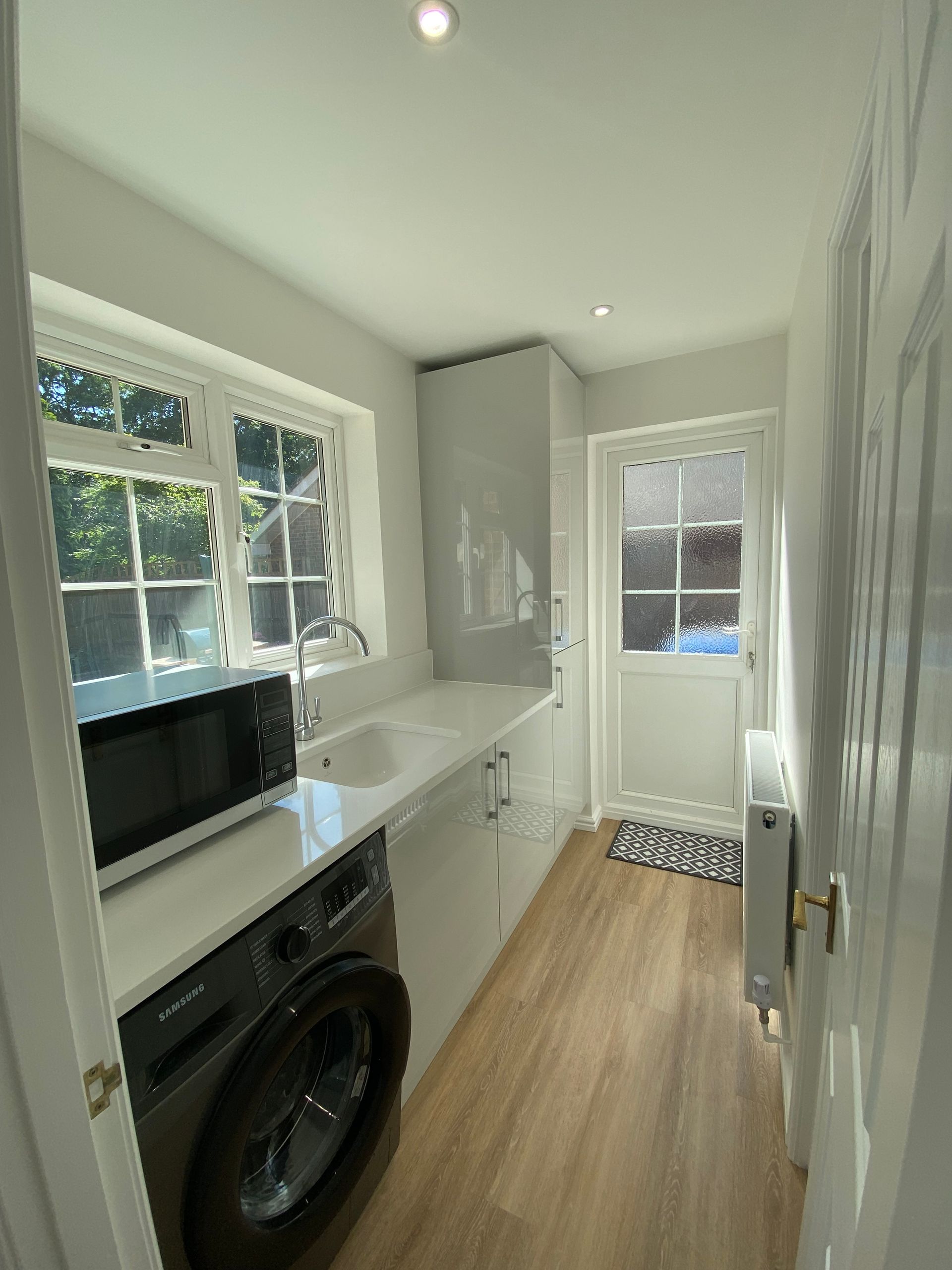 A laundry room with white cabinets, a light countertop, a gray washing machine, a microwave, and a back door.