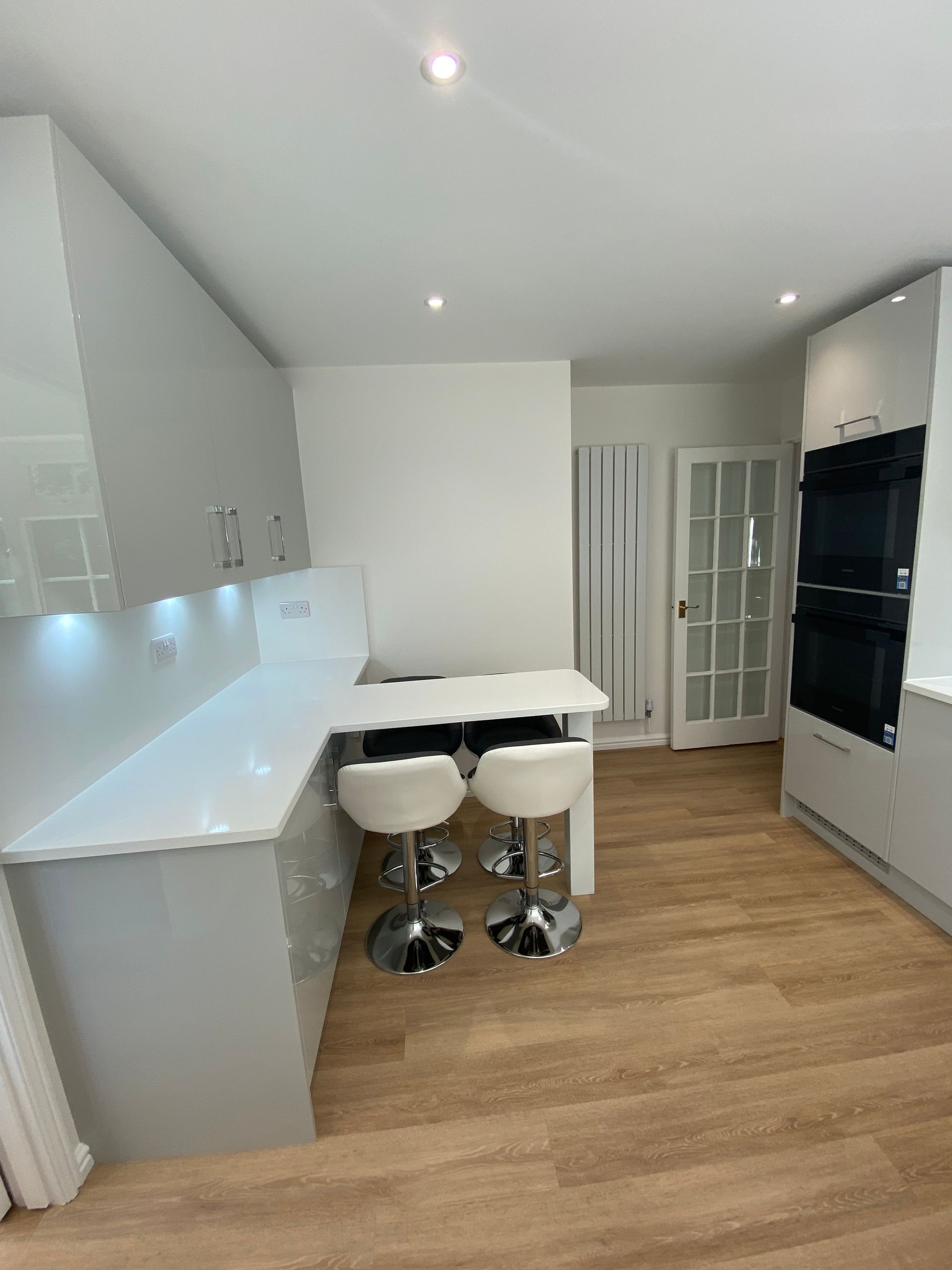 A modern kitchen with light grey cabinets, white countertops, a small dining table with two white stools, and black ovens.