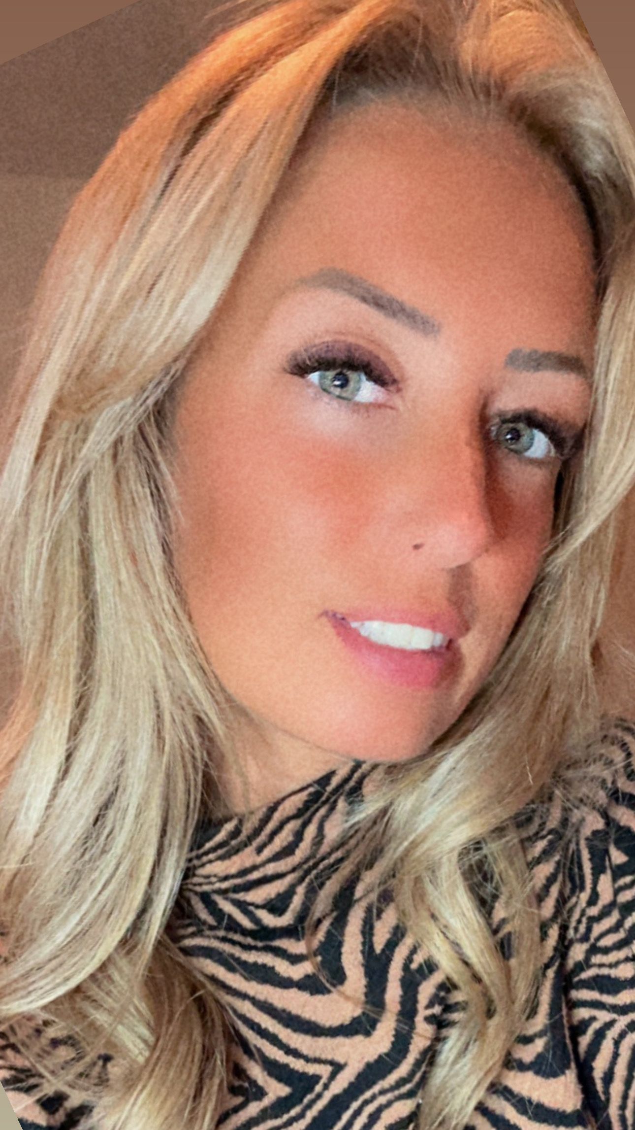 A close-up selfie of a person with blonde hair and blue eyes, wearing a brown and black zebra-print top.