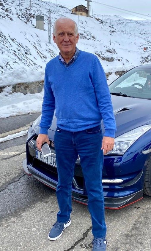 A person in a blue sweater and jeans standing outdoors in front of a dark blue sports car with snow-covered mountains.