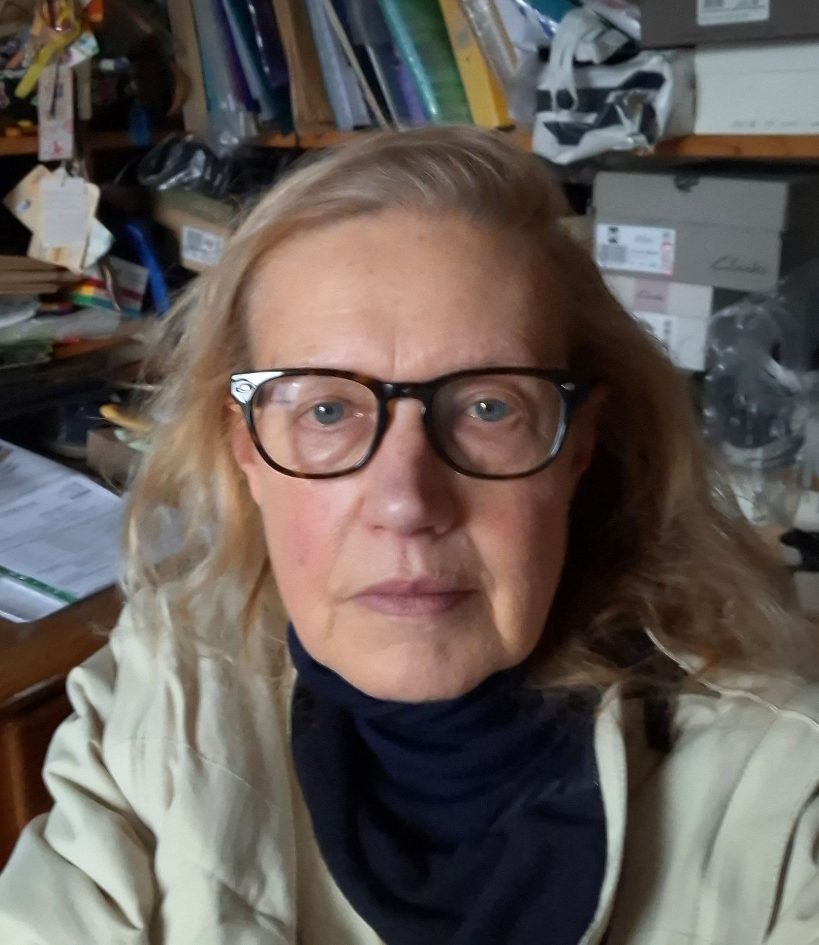 A person with long blonde hair and glasses, wearing a dark turtleneck and beige jacket, looking at the camera.