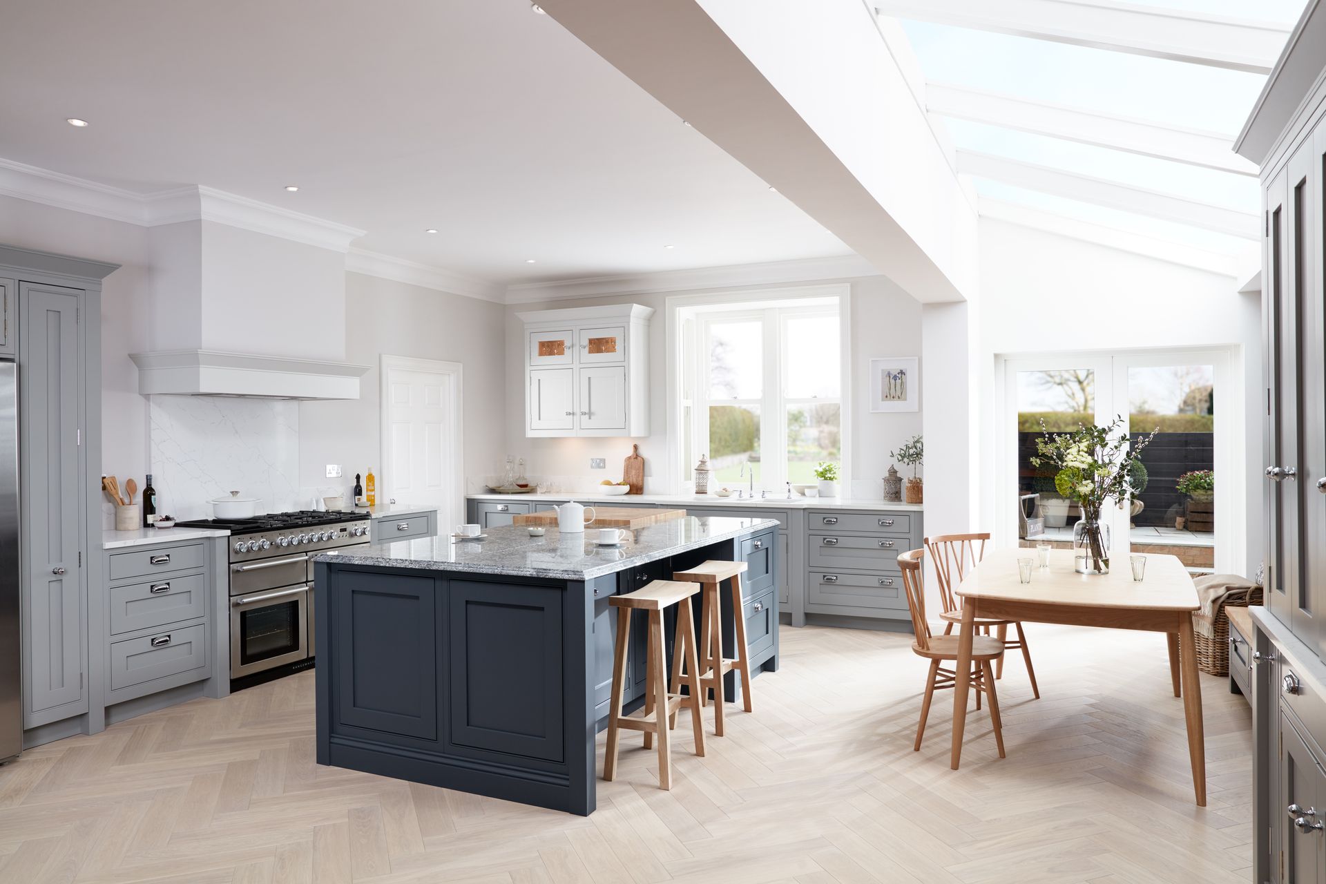 A modern, bright kitchen with blue-grey cabinets, a dark island, wooden stools, and a dining table under a skylight.