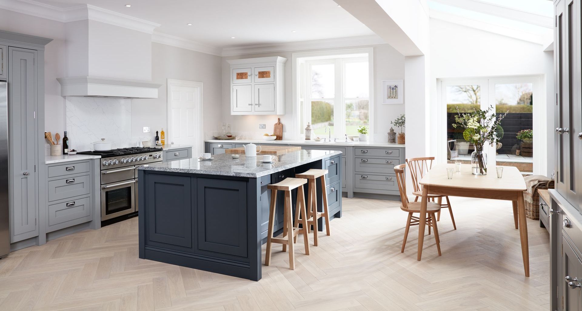A modern kitchen with grey cabinetry, a navy blue island, wooden stools, and a dining table on light wood herringbone floors.