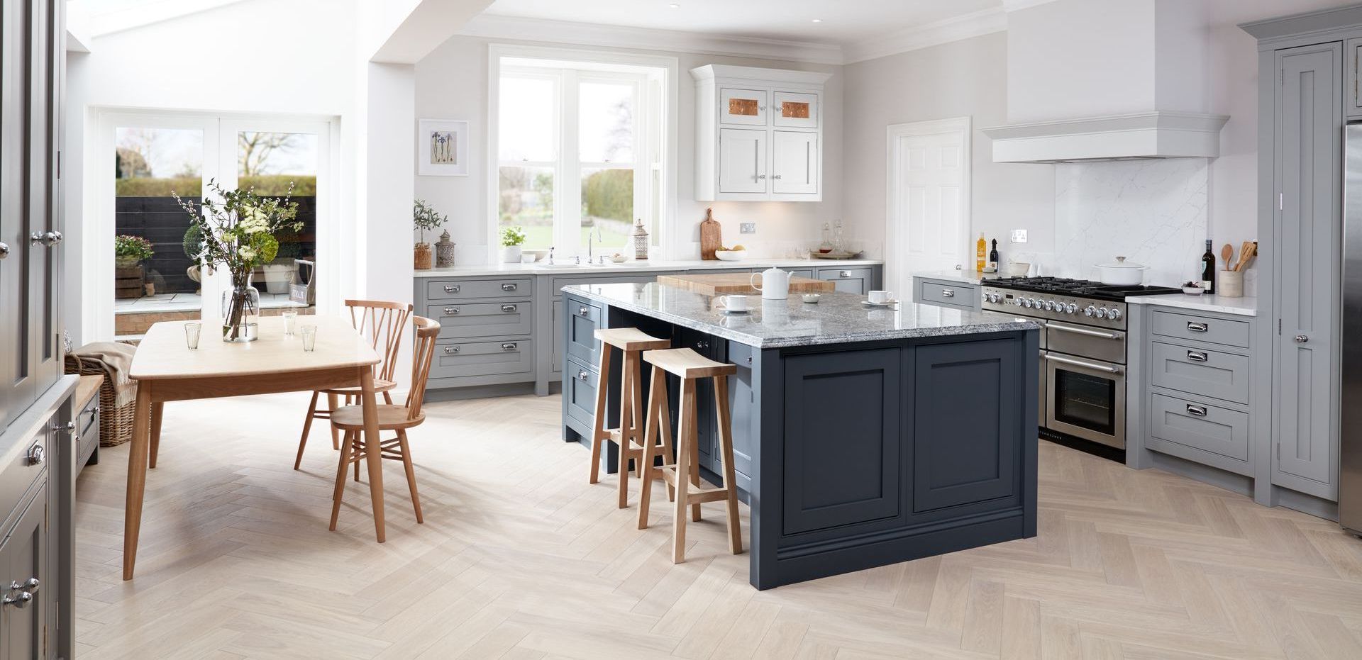 A spacious kitchen with a dark blue central island, light grey cabinets, wooden dining set, and herringbone wood floors.