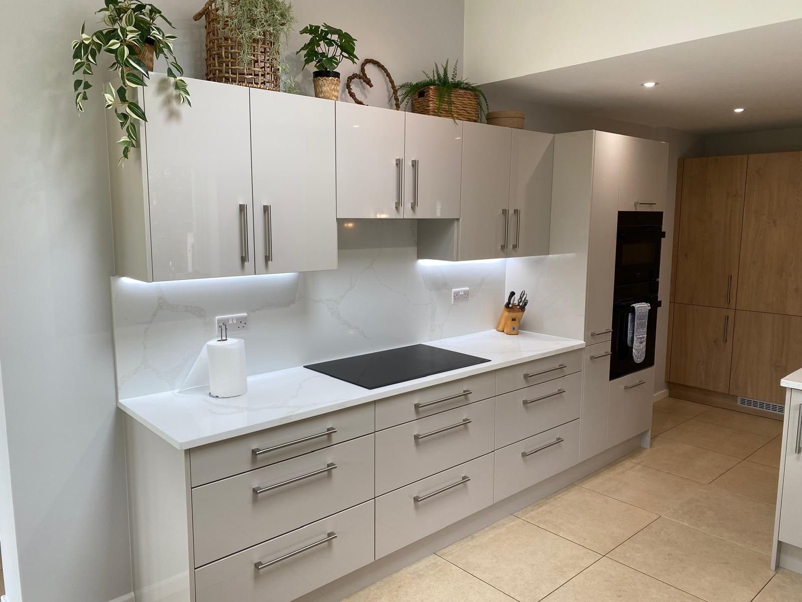 A sleek, modern kitchen with glossy off-white cabinets, a black induction cooktop, marble backsplash, and wooden accents.