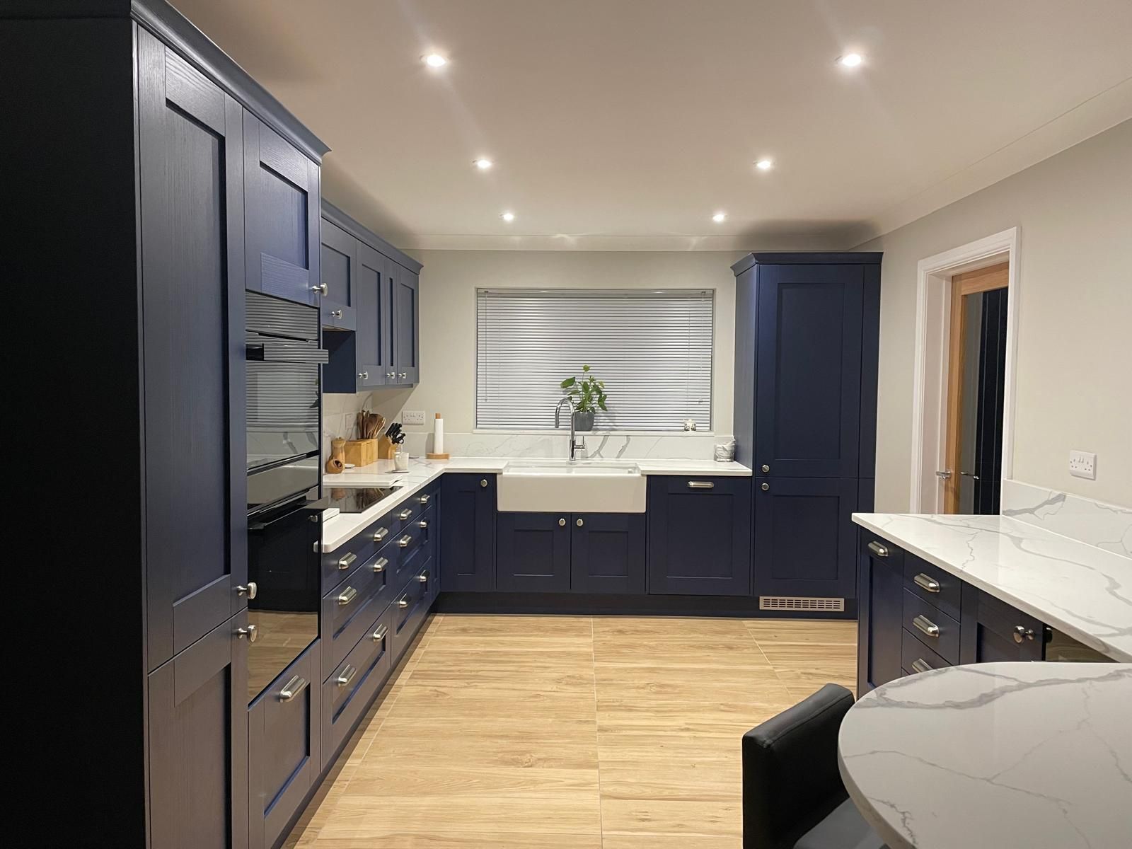 A modern kitchen with navy blue cabinets, white marble countertops, a farmhouse sink, and light wood-look tile flooring.